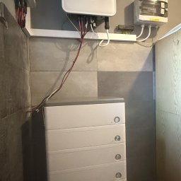 SUMID SPÓŁKA Z OGRANICZONĄ ODPOWIEDZIALNOŚCIĄ - Biały inwerter fotowoltaiczny marki FoxESS z podłączonymi kablami, zamontowany na ścianie nad modułową baterią energii w pomieszczeniu z szarą glazurą.