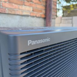 SUMID SPÓŁKA Z OGRANICZONĄ ODPOWIEDZIALNOŚCIĄ - Szary klimatyzator Panasonic na tle ceglanej ściany, widok z dołu, ukazujący detale obudowy i logo, z elementami zieleni w tle.
