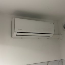 SUMID SPÓŁKA Z OGRANICZONĄ ODPOWIEDZIALNOŚCIĄ - Biały klimatyzator Rotenso zamontowany na ścianie w pomieszczeniu z marmurowymi płytkami. Widoczna instalacja odprowadzająca.