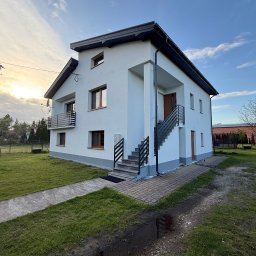 SUMID SPÓŁKA Z OGRANICZONĄ ODPOWIEDZIALNOŚCIĄ - Dwukondygnacyjny dom z białym tynkiem i szarą podmurówką, schody zewnętrzne z metalową balustradą, trawnik i fragment podjazdu. Widok z dołu, błękitne niebo.