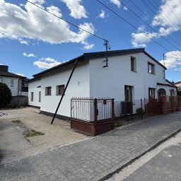 SUMID SPÓŁKA Z OGRANICZONĄ ODPOWIEDZIALNOŚCIĄ - Biały dom jednorodzinny z brązowymi oknami i ogrodzeniem, widziany z chodnika. Błękitne niebo z chmurami w tle. Widoczne podpory elewacji.