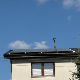 SUMID SPÓŁKA Z OGRANICZONĄ ODPOWIEDZIALNOŚCIĄ - Panele słoneczne na dachu domu jednorodzinnego z beżową elewacją i brązowymi oknami, na tle błękitnego nieba z delikatnymi obłokami.