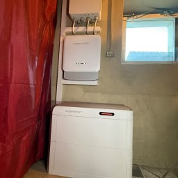 SUMID SPÓŁKA Z OGRANICZONĄ ODPOWIEDZIALNOŚCIĄ - Biały magazyn energii Sigenenergy zamontowany na ścianie w pomieszczeniu z oknem i zasłoną. Widoczne kable i instalacja elektryczna.