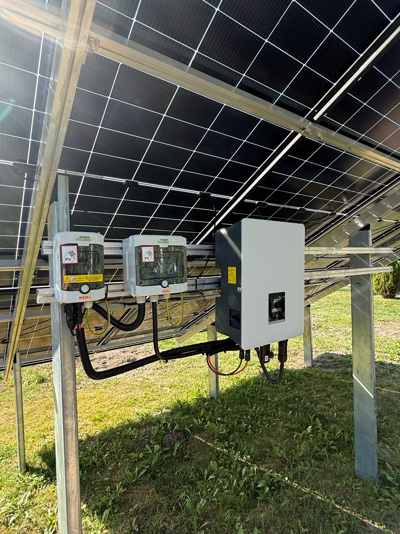 Instalacja fotowoltaiczna: widok od spodu na panele, inwerter FOX-O i dwa ograniczniki przepięć Noark Keno, zamontowane na konstrukcji wsporczej na trawniku.