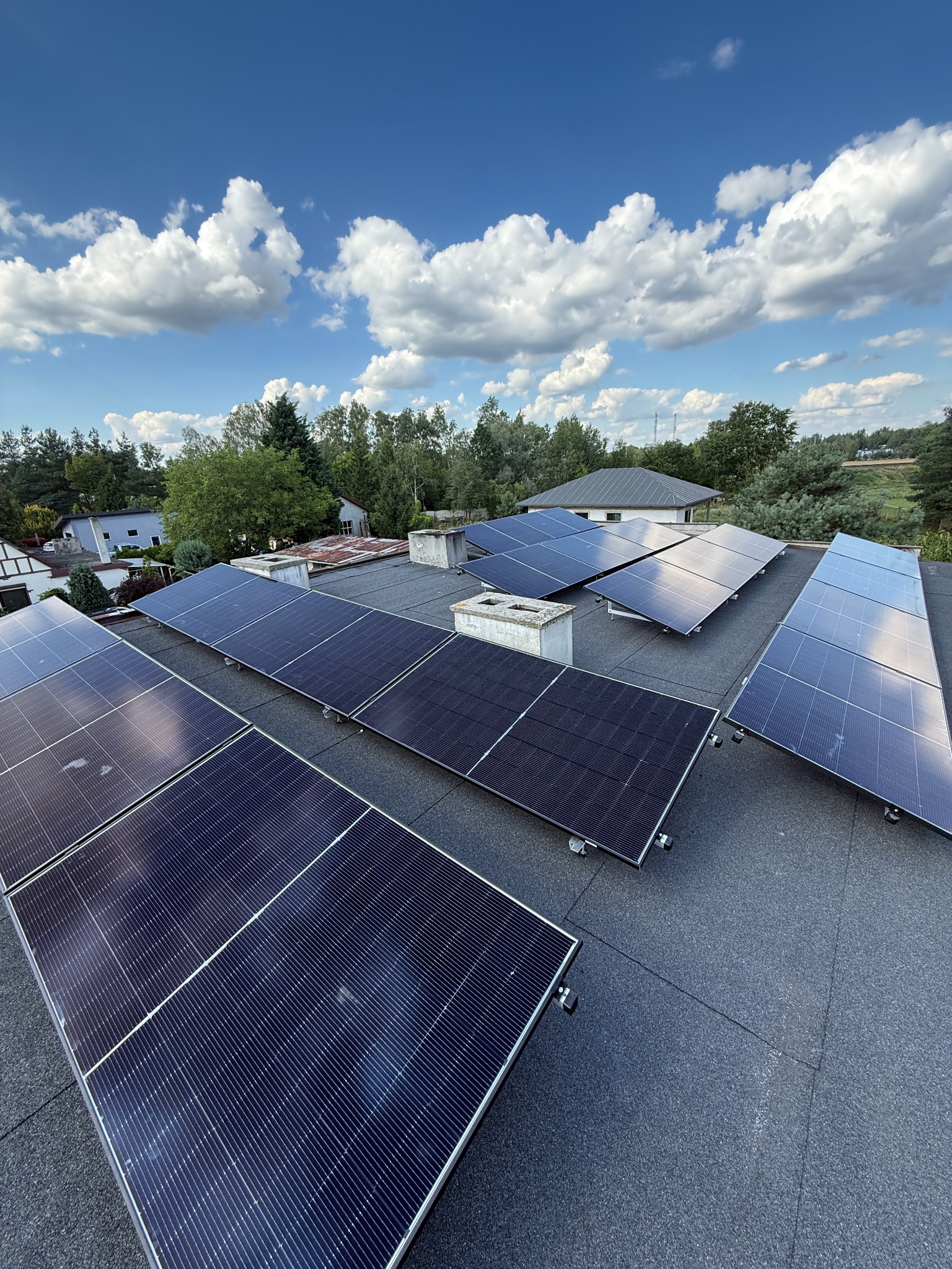 Instalacja fotowoltaiczna na dachu budynku w słoneczny dzień, widok z góry. Panele skierowane w stronę nieba z chmurami. Widoczne domy w tle.