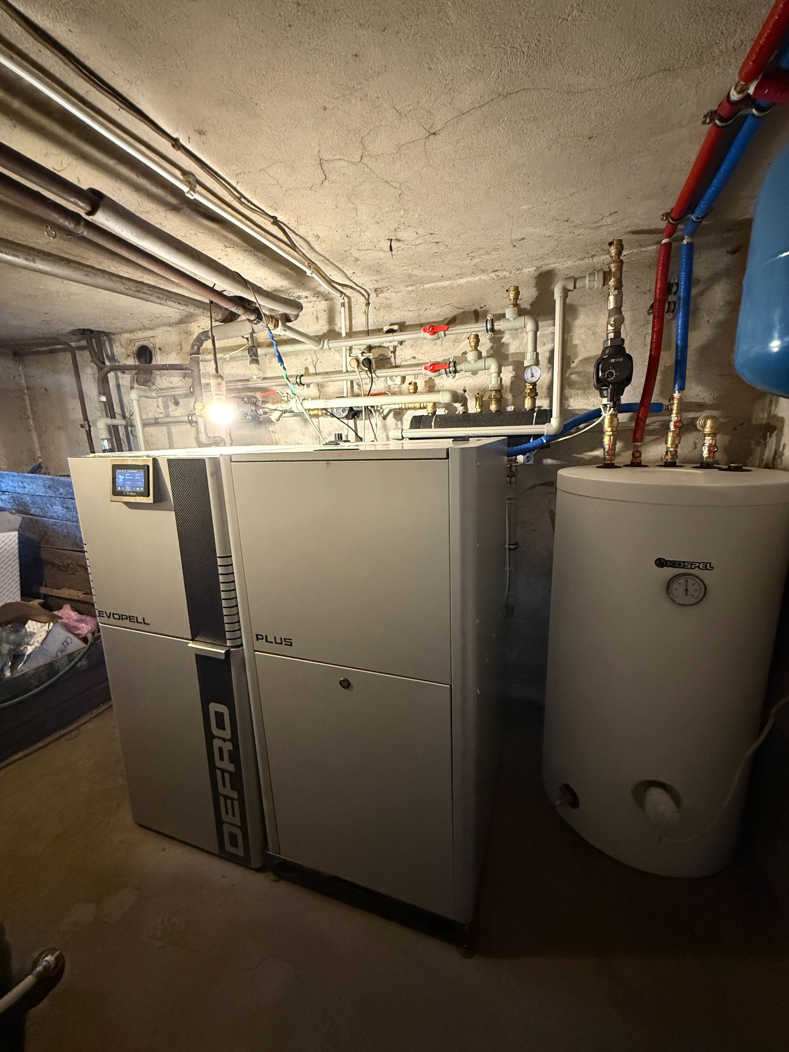 Nowoczesny piec Defro Evopell Plus z zbiornikiem Kospel Concept w kotłowni. Widoczne rury instalacji grzewczej i oświetlenie sufitowe.