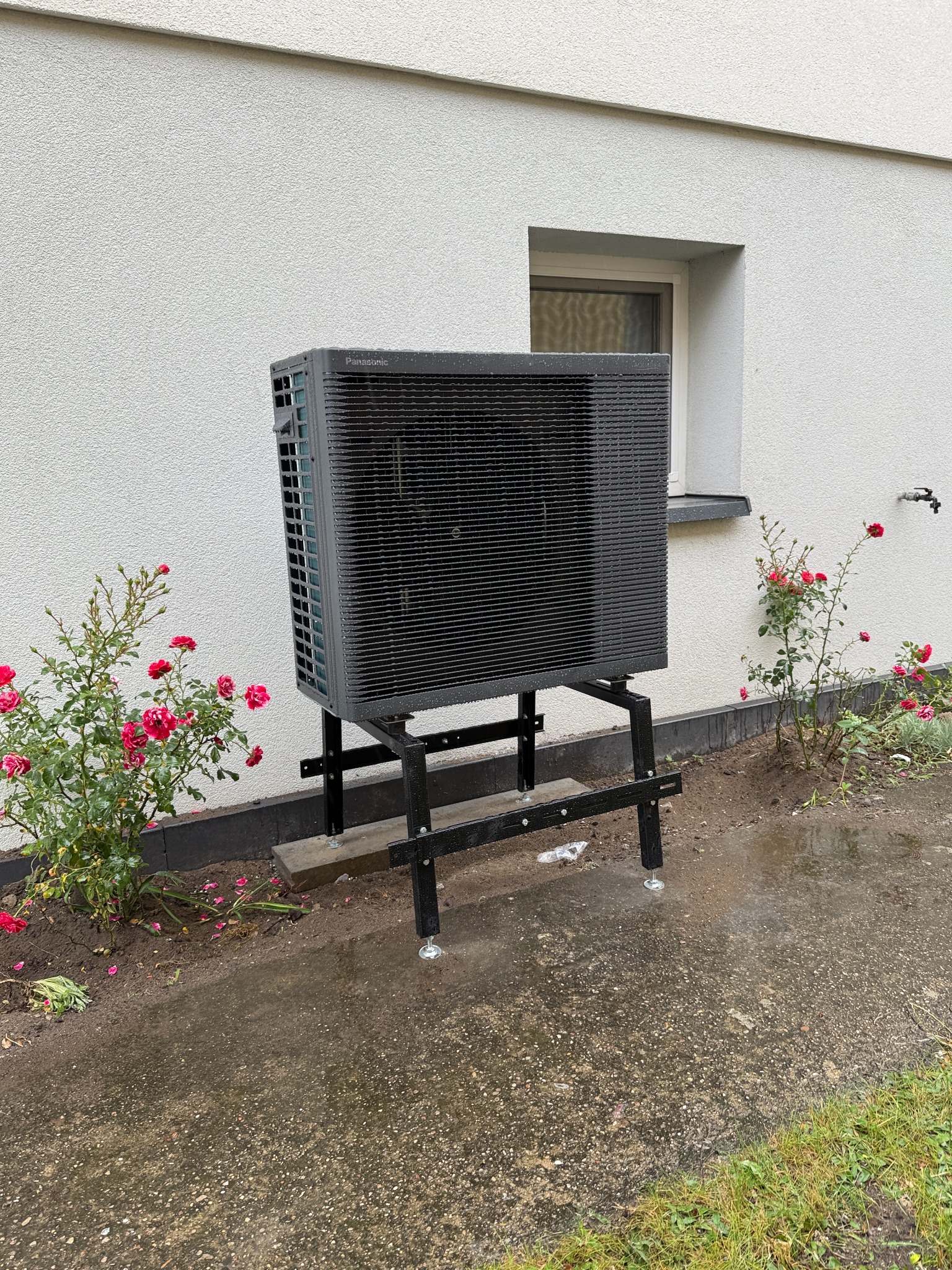 Zewnętrzna jednostka pompy ciepła Panasonic na metalowym stelażu, ustawiona przy ścianie budynku z oknem i rabatą róż. Widoczne ślady deszczu na podłożu.