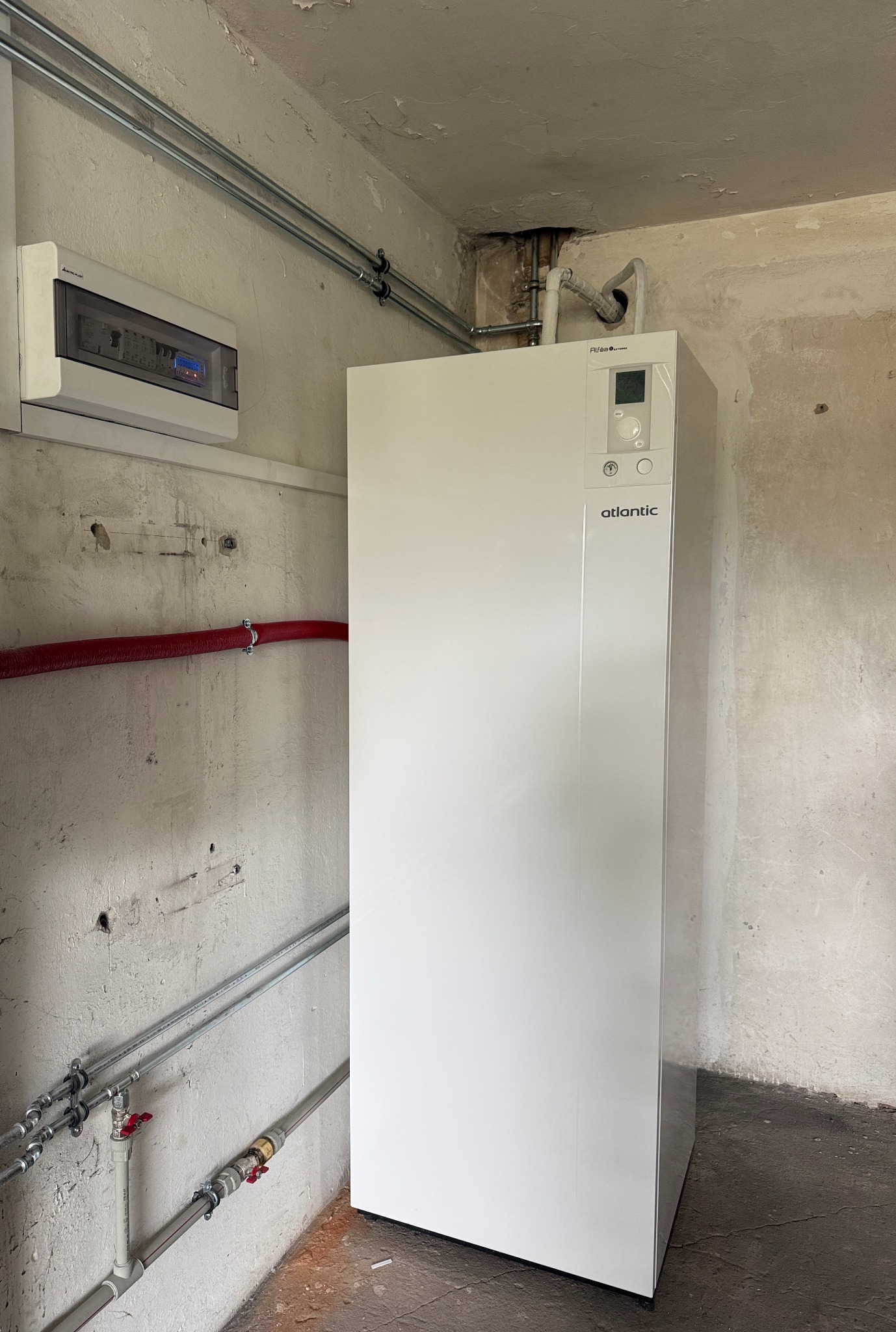 Biała pompa ciepła Atlantic w surowym pomieszczeniu z instalacją rurową i skrzynką elektryczną na ścianie. Widoczne ślady niedokończonych prac budowlanych.