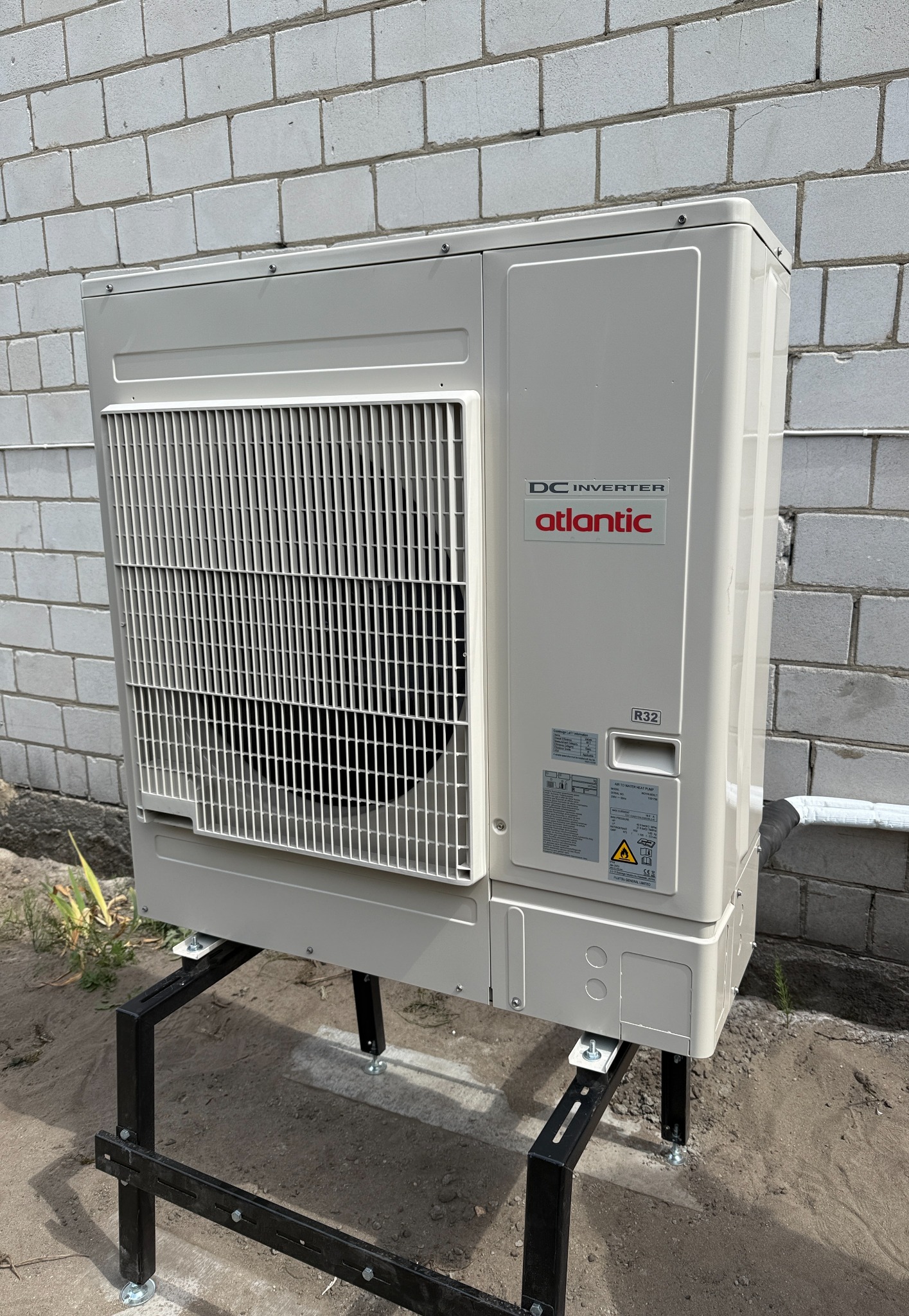 Zewnętrzna jednostka pompy ciepła Atlantic, zamontowana na czarnej, metalowej konstrukcji przy ścianie z białego bloczka. Widoczny model DC Inverter R32.