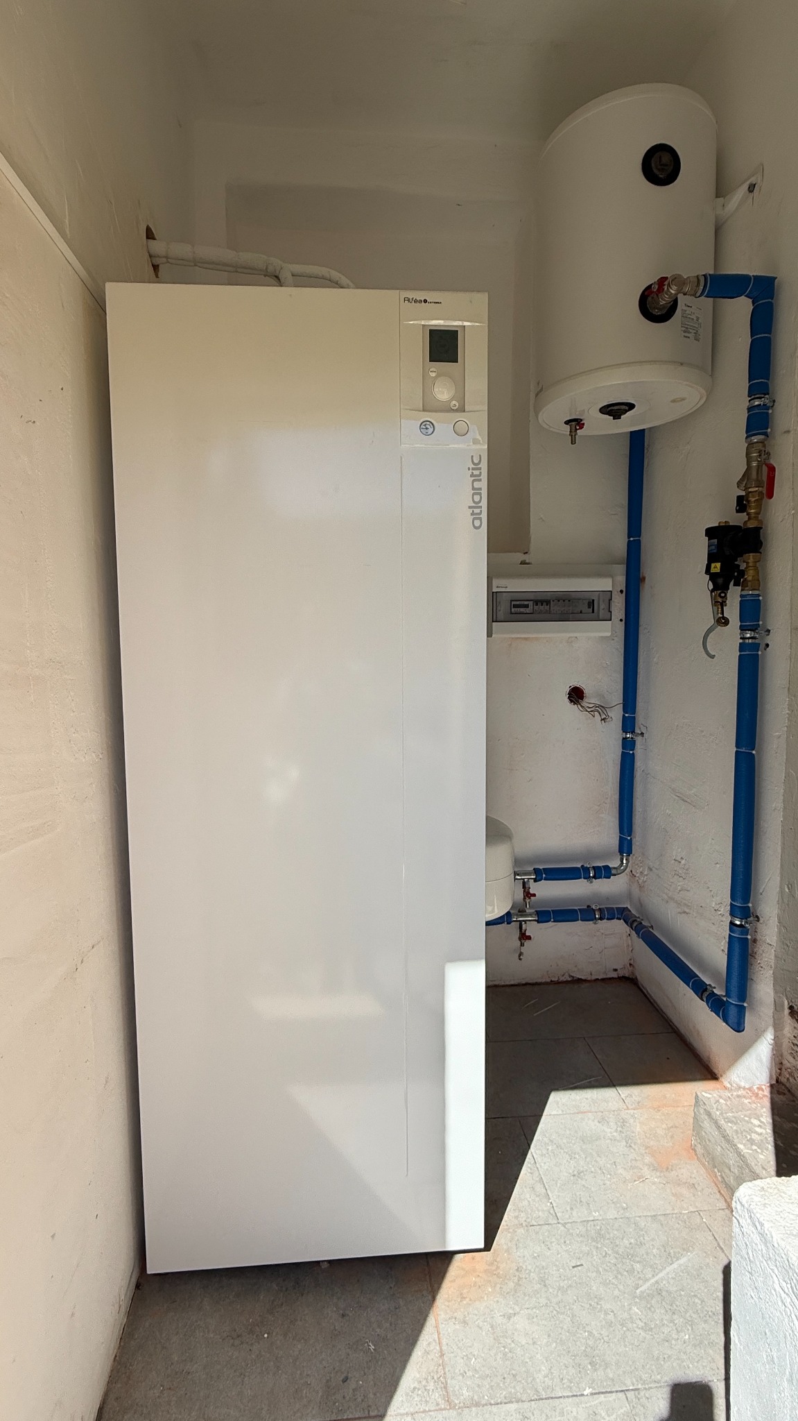 Biała pompa ciepła Atlantic z zasobnikiem wody, zainstalowana w pomieszczeniu z niebieskimi rurami i szarą podłogą. Widoczna skrzynka elektryczna i fragment ściany.