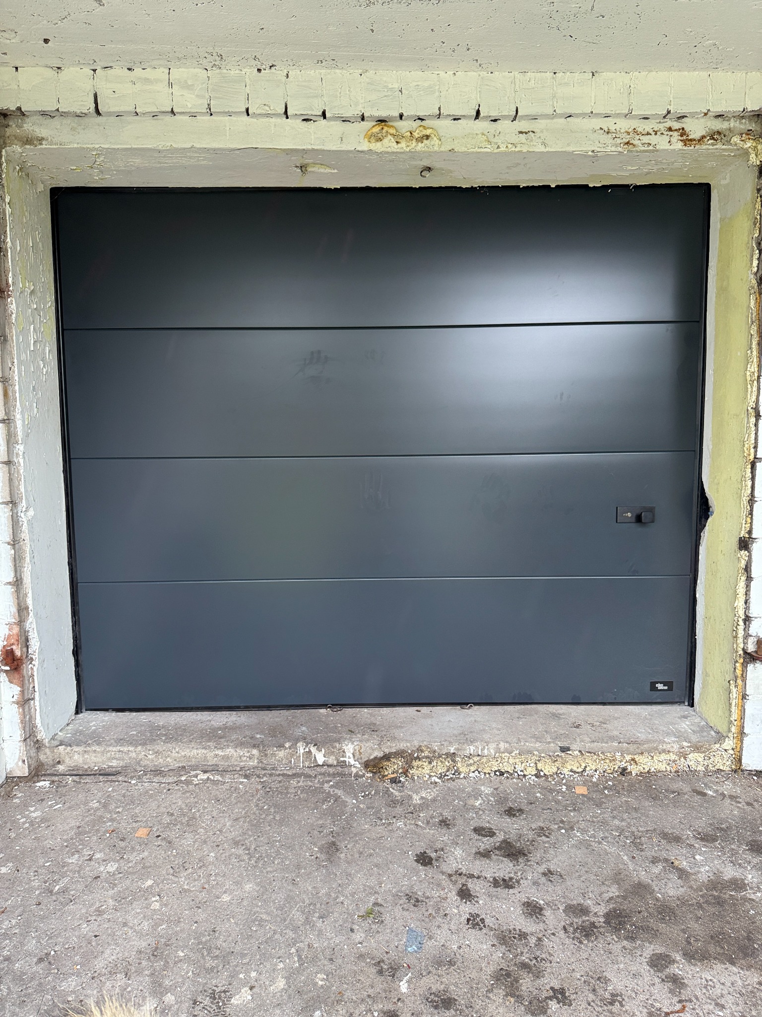 Nowa, antracytowa brama garażowa segmentowa w starej, odrapanej ramie z cegły i betonu. Widoczne trzy poziome segmenty i czarny uchwyt.