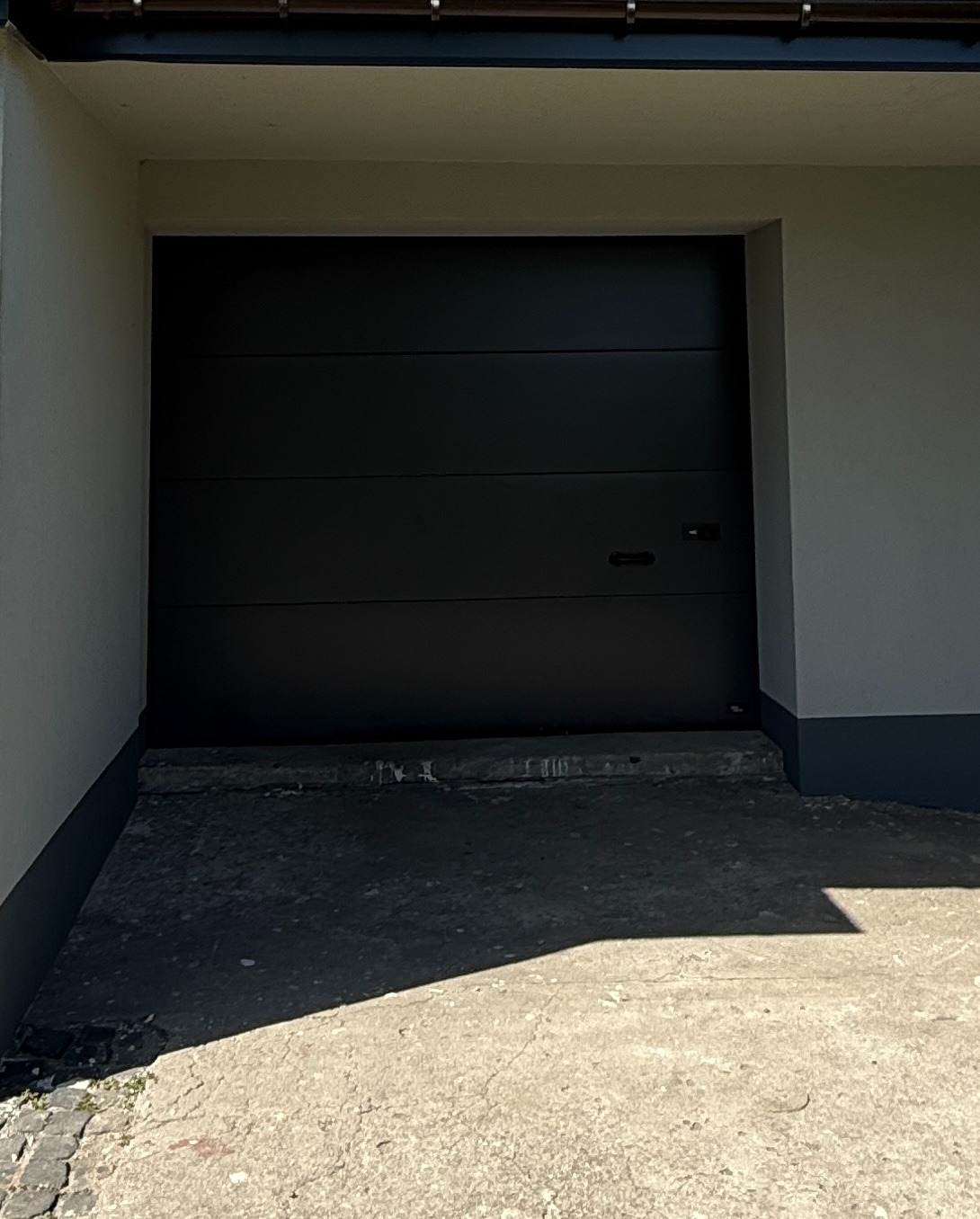 Zamknięta, grafitowa brama garażowa z poziomymi panelami, zamontowana w jasnym budynku. Widoczny betonowy podjazd i fragment orynnowania.