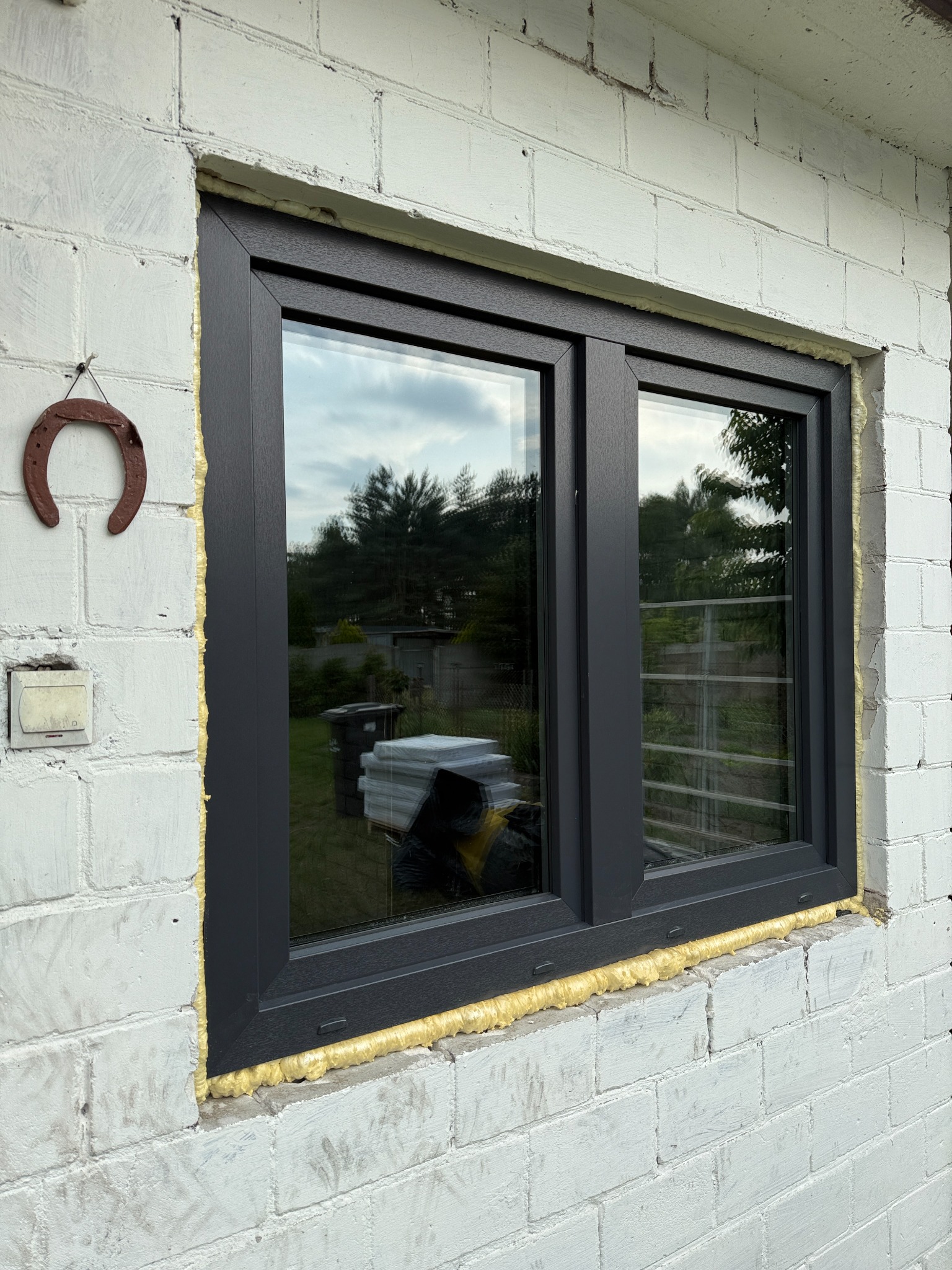 Szare okno PCV zamontowane w białej ścianie z pustaków, uszczelnione pianką montażową. Na ścianie wisi podkowa. W szybie okna odbija się zieleń drzew.