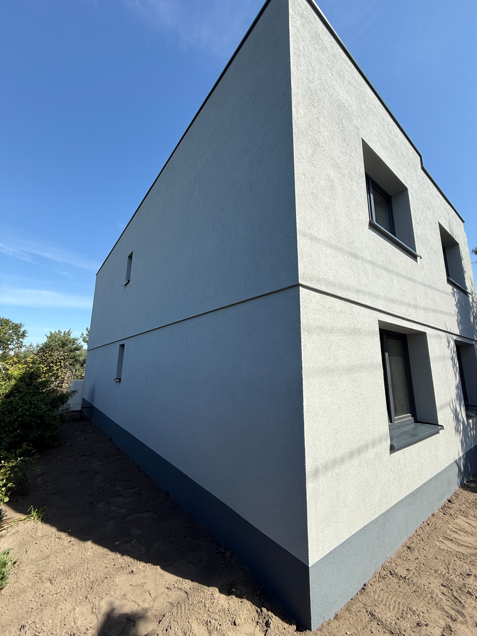 Dwukondygnacyjny dom z oknami i świeżo otynkowaną, szarą elewacją, widoczny z perspektywy żabiej, na tle błękitnego nieba. Uwagę zwraca minimalistyczny design.