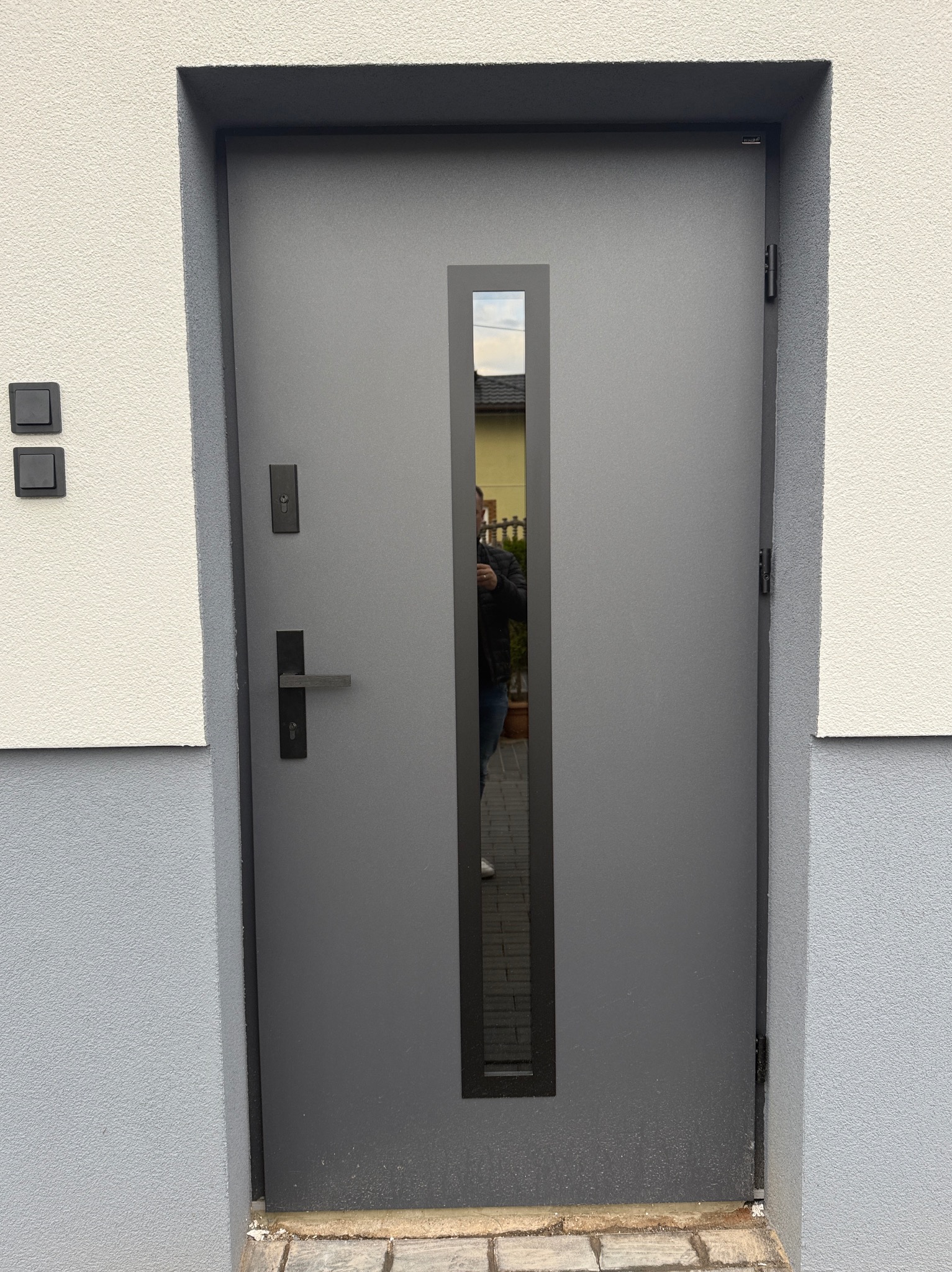 Szare, nowoczesne drzwi wejściowe z pionowym przeszkleniem, wkomponowane w elewację budynku o jasnej, strukturalnej powierzchni i szarym cokole. Widoczne czarne włączniki światła.