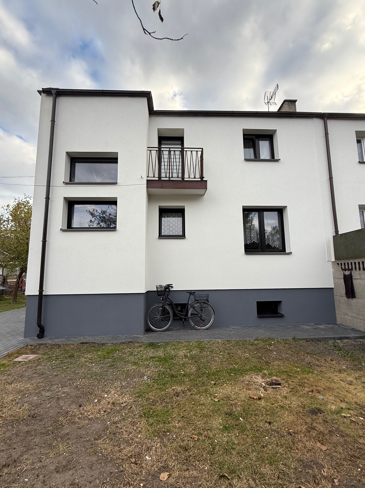 Dwupiętrowy dom z białymi ścianami i ciemnymi oknami. Na parterze szara lamperia. Opiera się o nią rower z koszykiem. U góry antena.