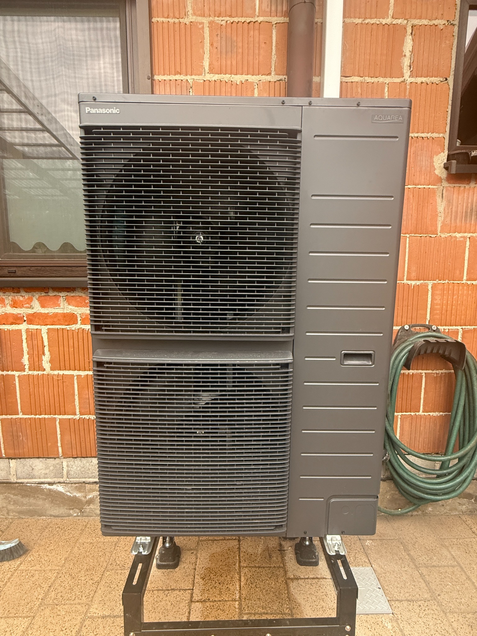 Szara pompa ciepła Panasonic Aquarea na zewnątrz, ustawiona na metalowym stelażu, z widoczną fasadą z cegły i fragmentem okna w tle.