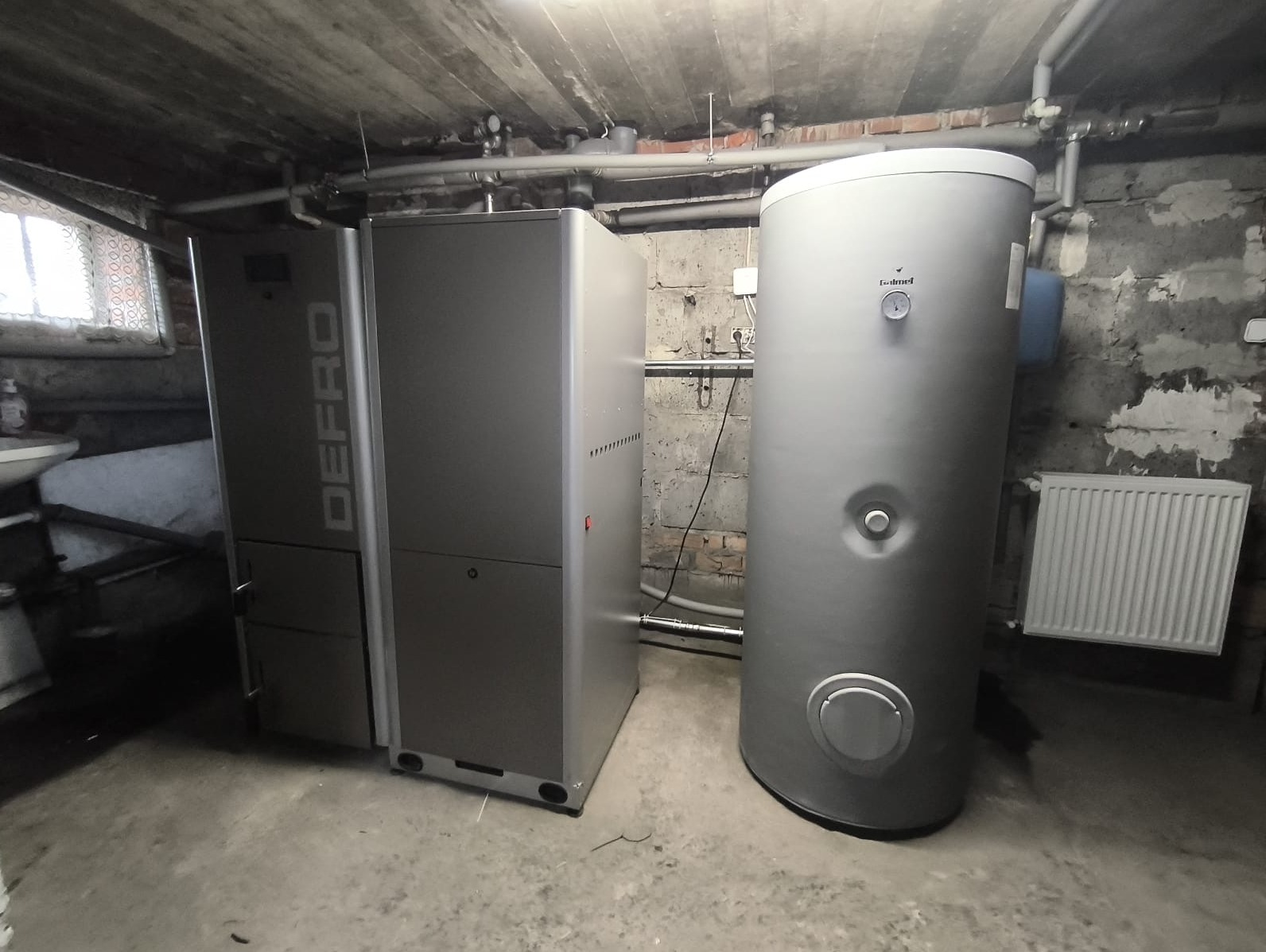 Szary piec Defro i zasobnik ciepłej wody w kotłowni z surowym betonowym sufitem. Widoczna instalacja rurowa i grzejnik. Ujęcie z niskiego kąta.