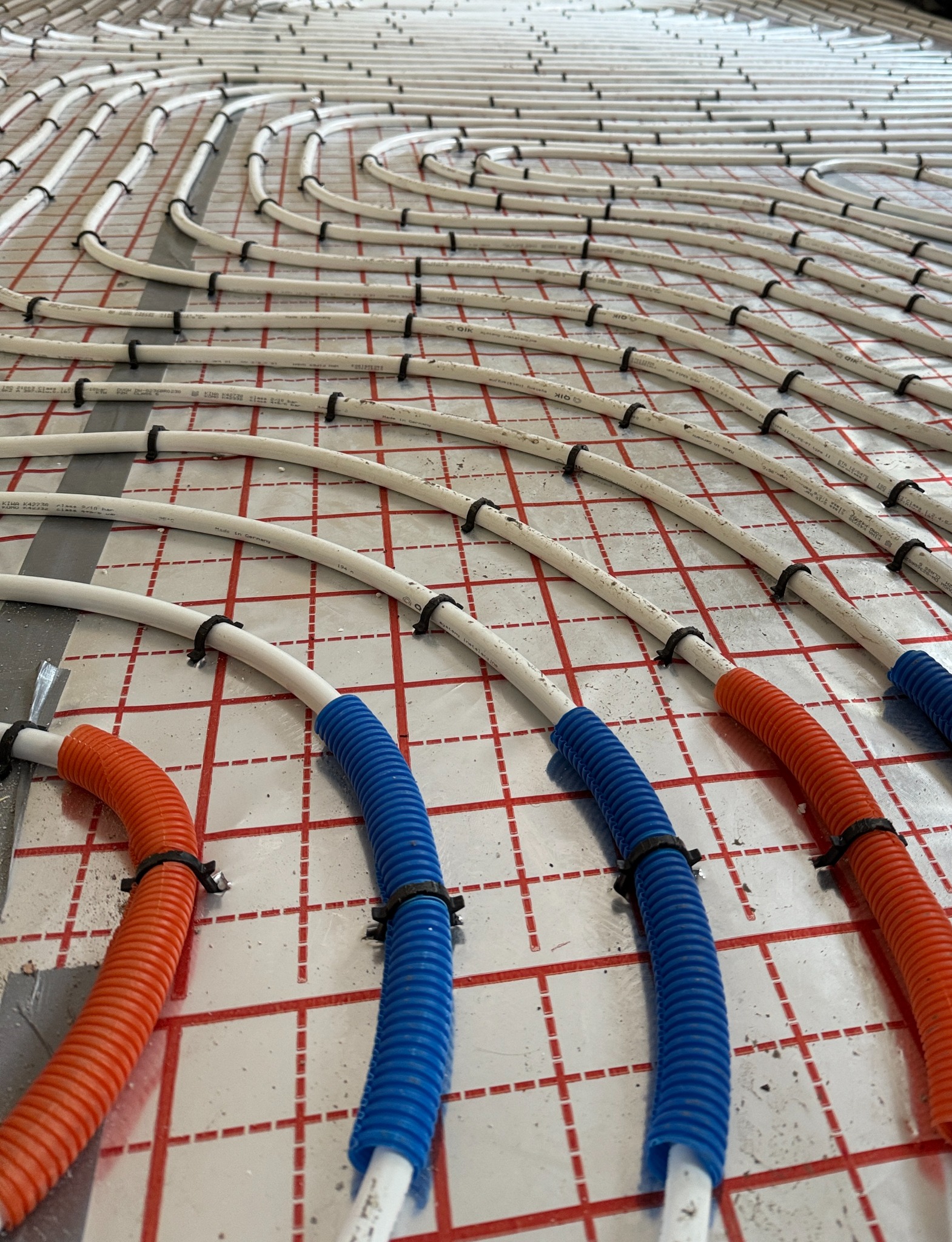 Rozłożona instalacja ogrzewania podłogowego z rurami w kolorze białym, pomarańczowym i niebieskim, przymocowanymi czarnymi uchwytami do podłoża z siatką.