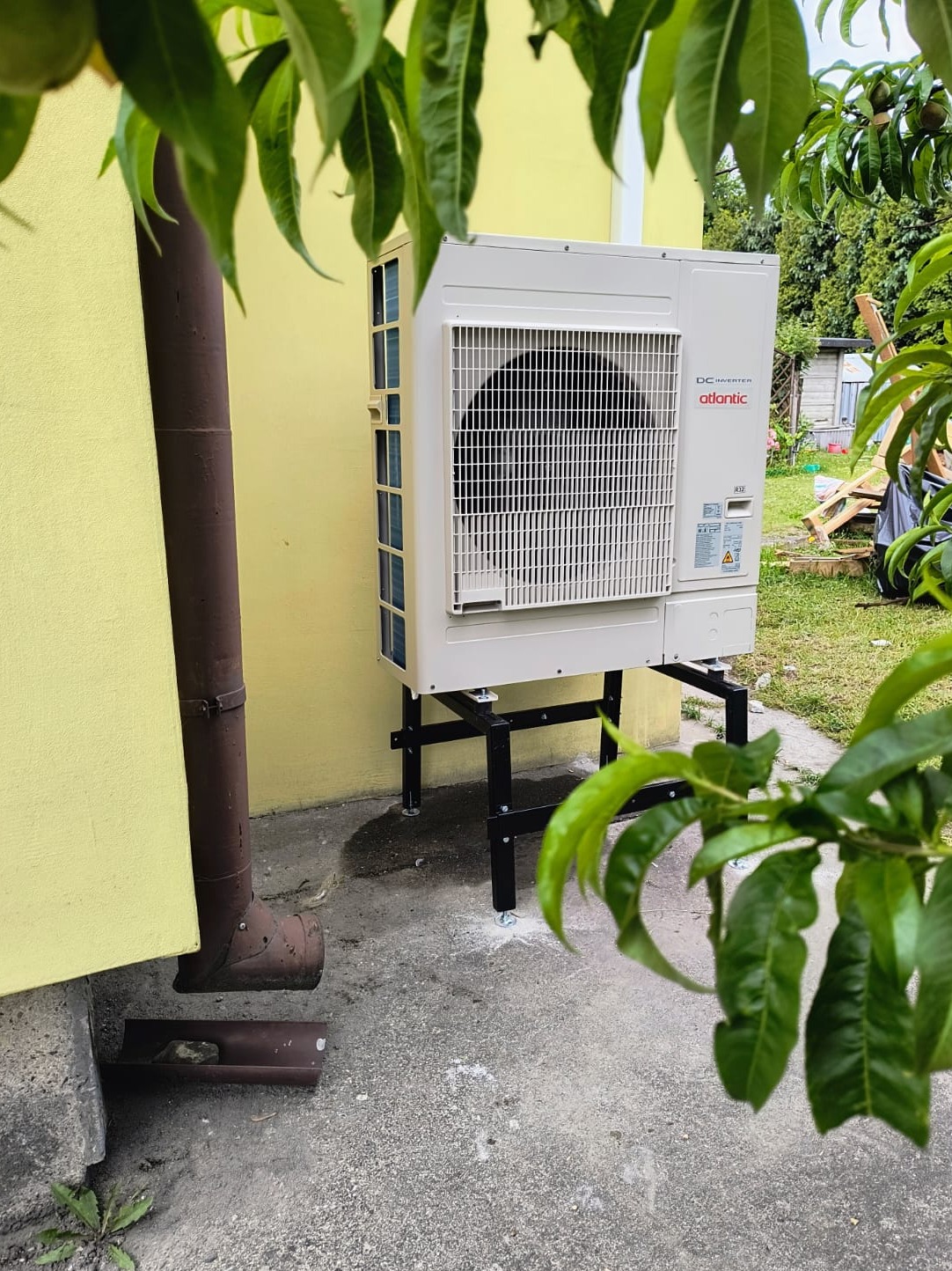 Biała pompa ciepła Atlantic DC Inverter zamontowana na czarnym stelażu przy żółtej ścianie budynku, widoczna zza gałęzi drzewa owocowego.