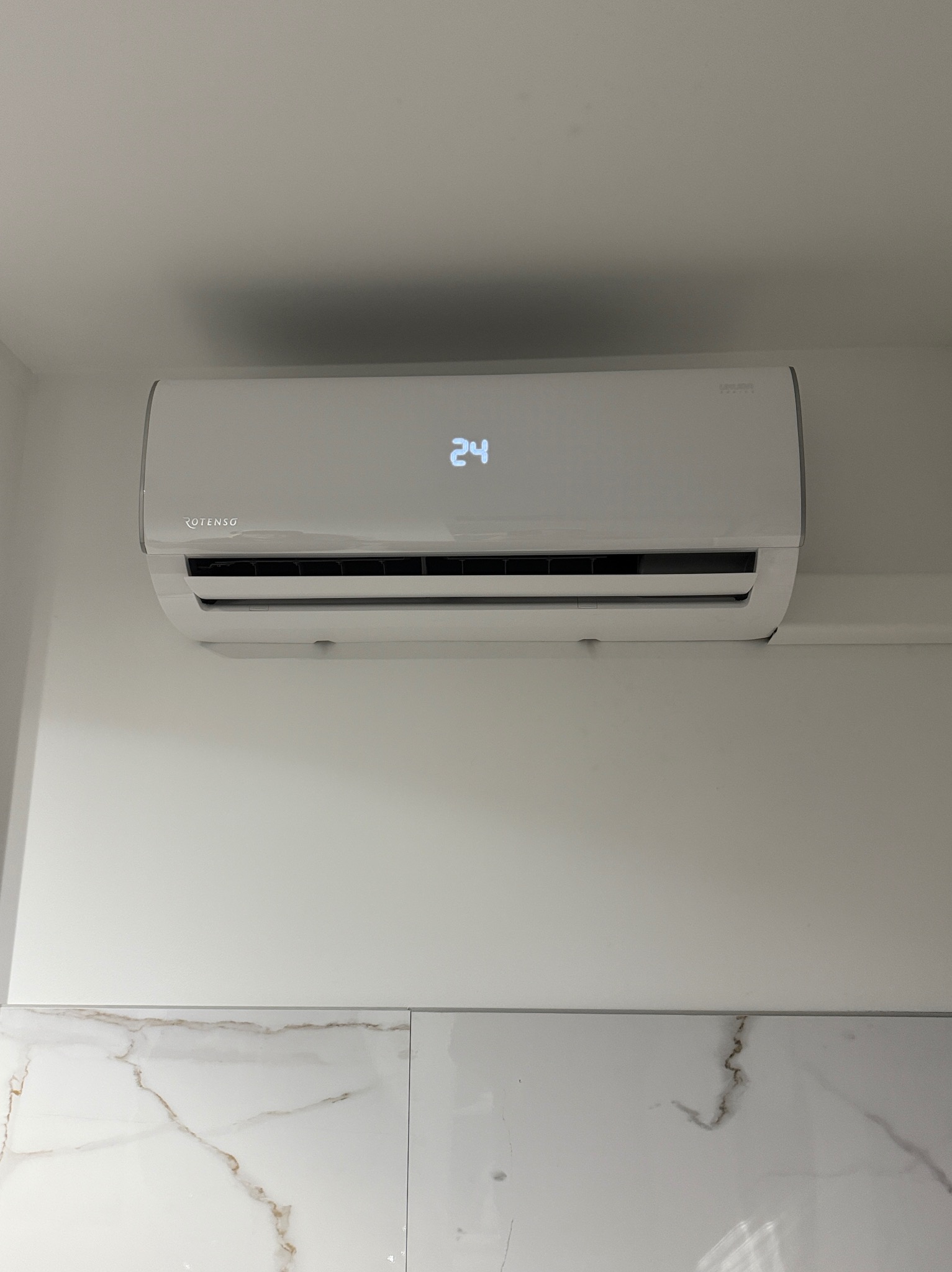 Biały klimatyzator Rotenso z wyświetlaczem 24°C, zamontowany na białej ścianie nad marmurowymi płytkami. Minimalistyczny design, czysty montaż.