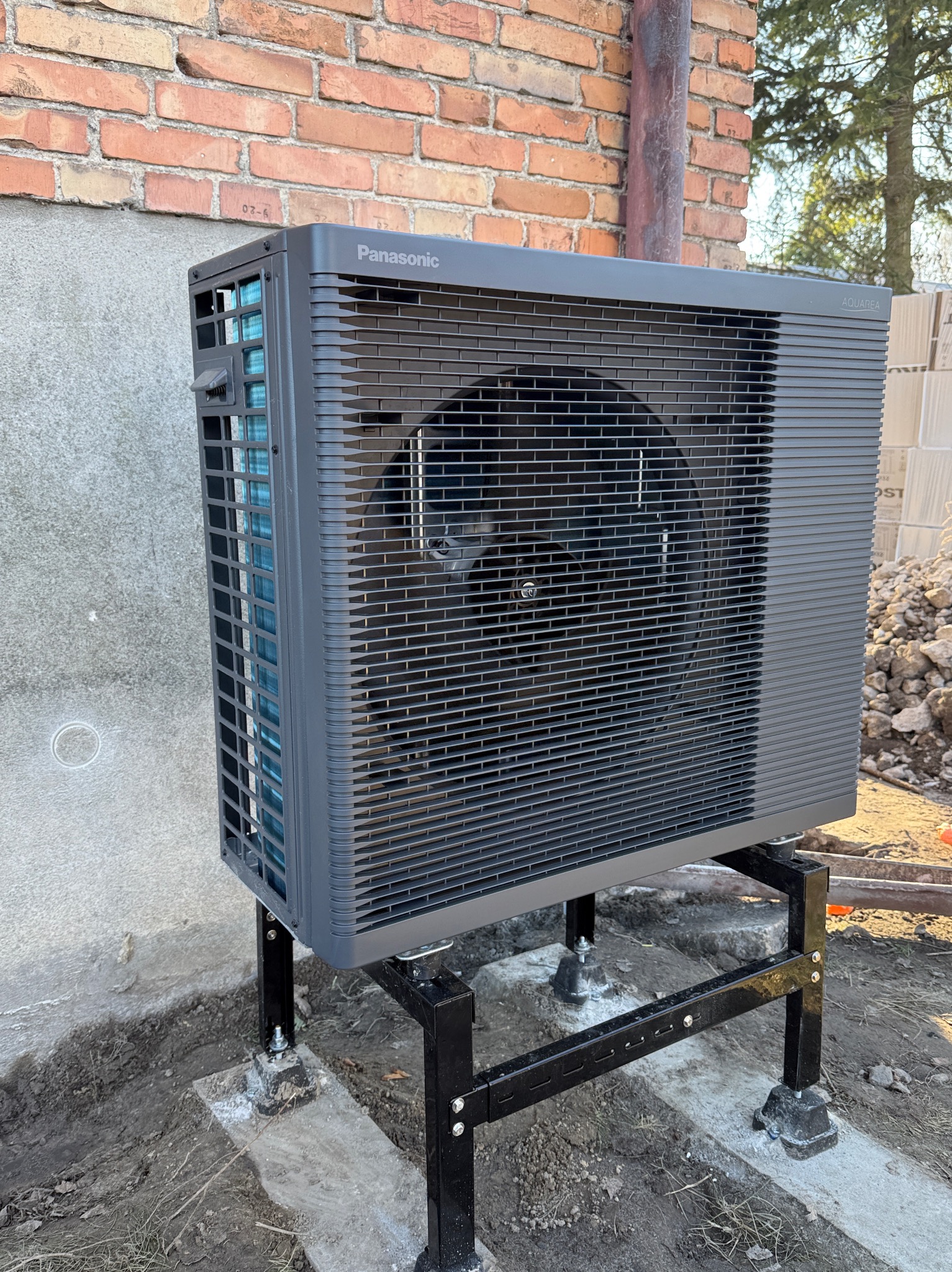 Szara pompa ciepła Panasonic Aquarea, zamontowana na czarnej, metalowej konstrukcji, na tle ceglanej ściany i ziemi. Widoczne elementy instalacji.