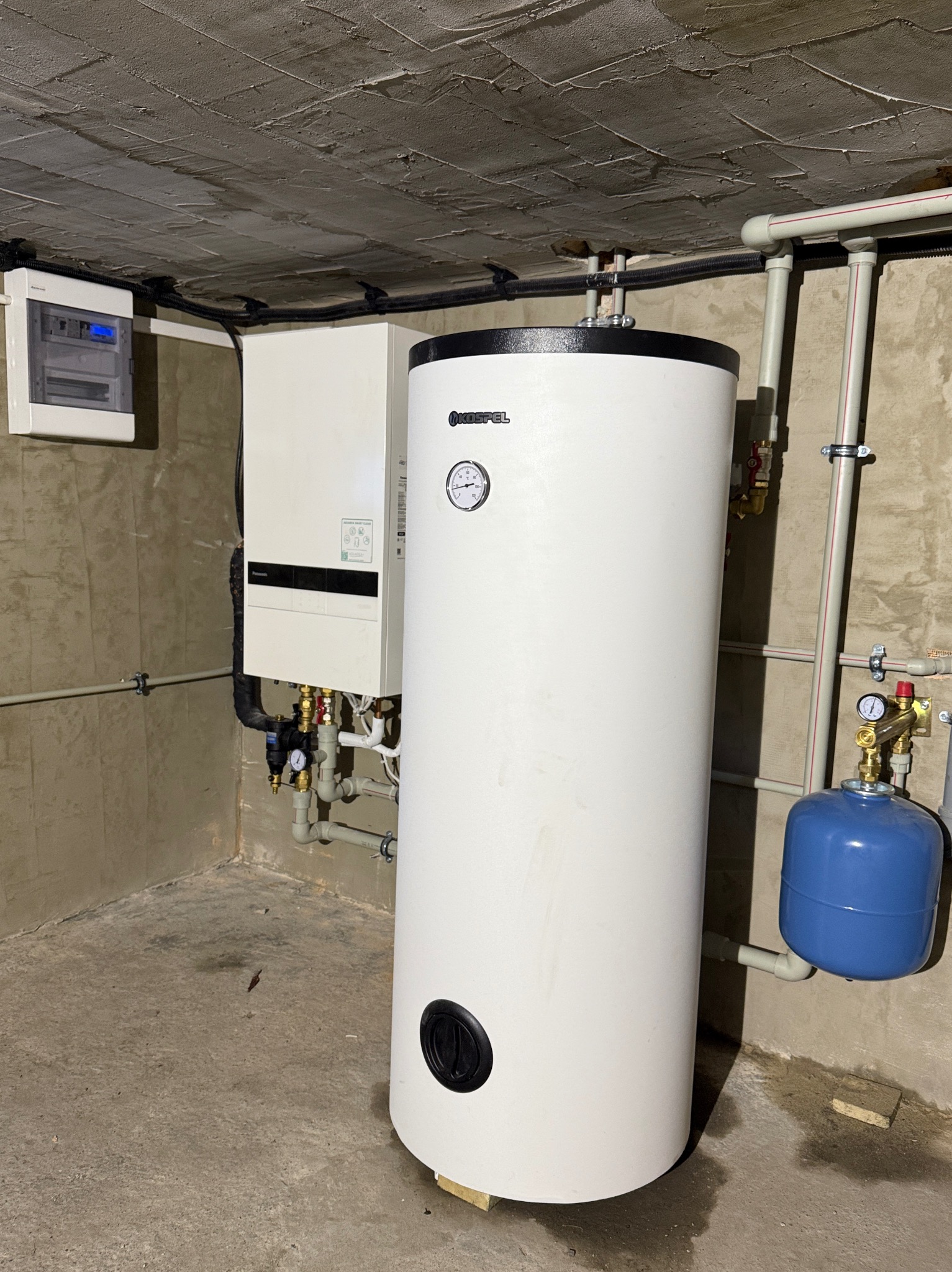 Biały, cylindryczny zasobnik ciepłej wody marki Kospel z manometrem, obok kotła grzewczego Panasonic, w surowym pomieszczeniu z betonowym sufitem i instalacją.