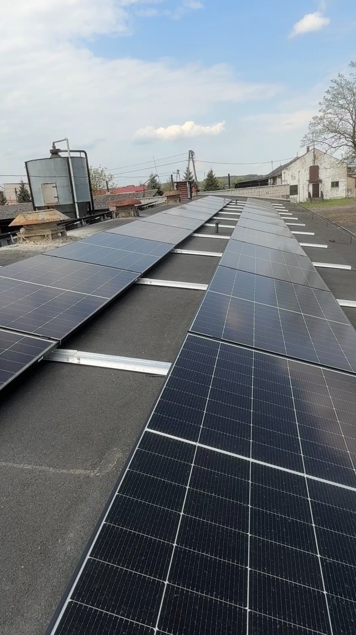 Rząd paneli fotowoltaicznych na płaskim dachu budynku gospodarczego, widok z góry, błękitne niebo w tle. Montaż paneli na konstrukcji wsporczej.