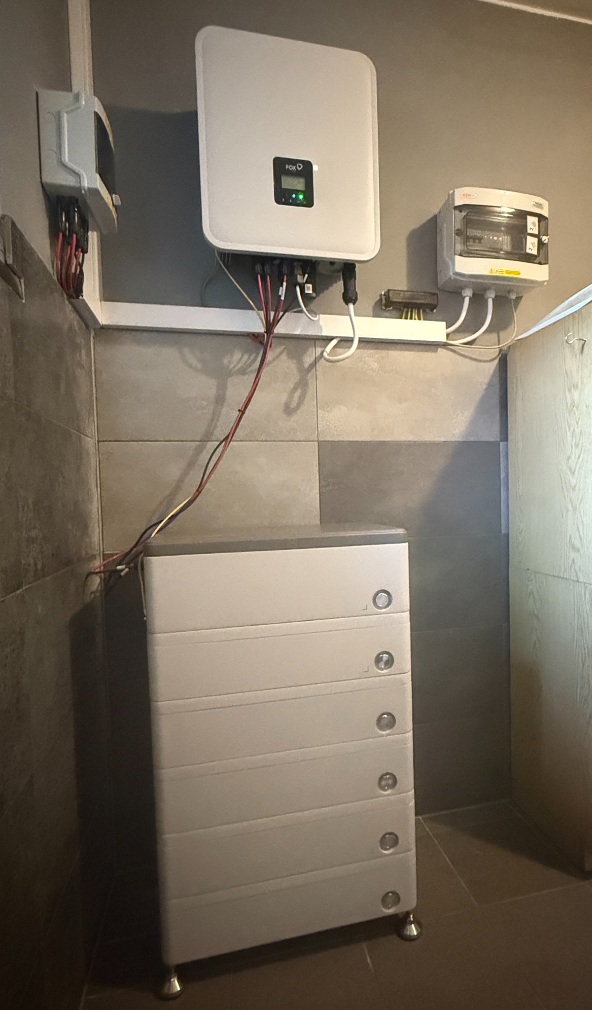 Biały inwerter fotowoltaiczny marki FoxESS z podłączonymi kablami, zamontowany na ścianie nad modułową baterią energii w pomieszczeniu z szarą glazurą.