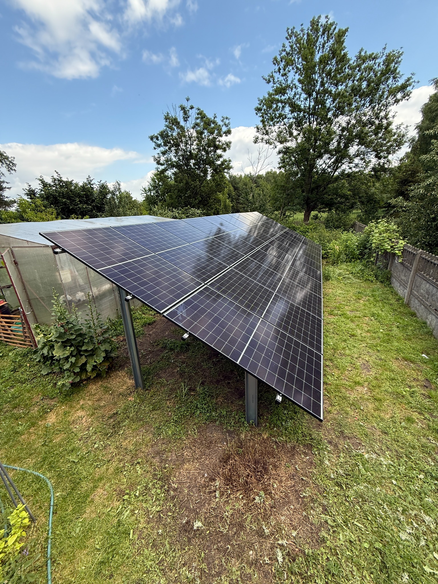 Panel fotowoltaiczny na metalowej konstrukcji, zainstalowany na trawniku obok szklarni i betonowego ogrodzenia w słoneczny dzień. Widok z góry.