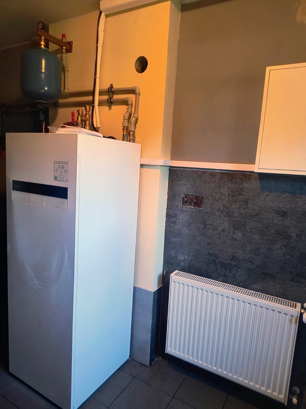 Biała pompa ciepła Panasonic Aquarea z niebieskim zbiornikiem wyrównawczym, rurami i grzejnikiem w pomieszczeniu z szaro-żółtymi ścianami i ciemną płytką.