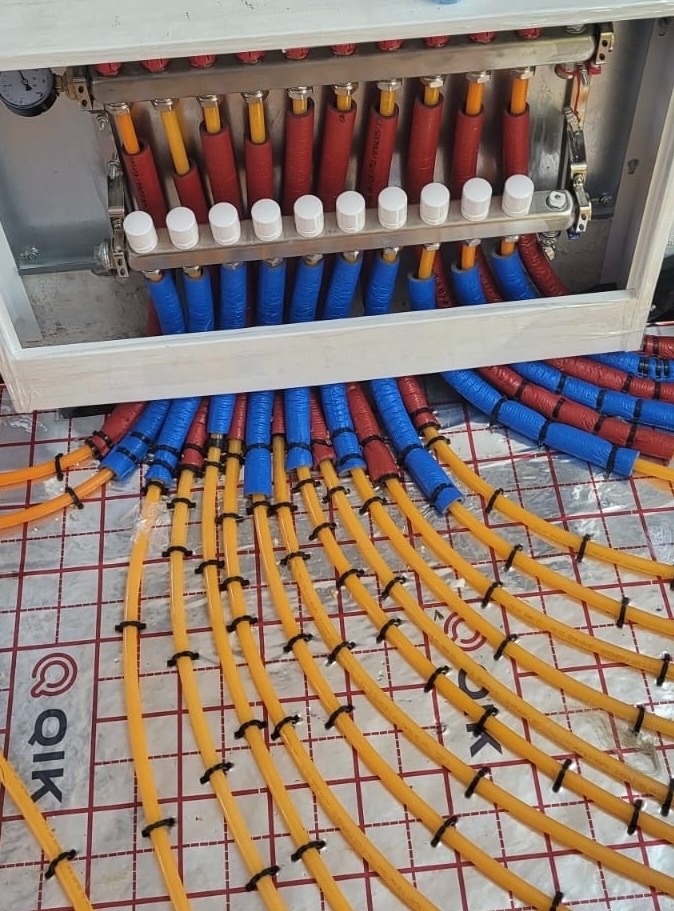 Rozdzielacz ogrzewania podłogowego z rurami w izolacji (czerwonej i niebieskiej) ułożonymi na folii z nadrukowaną siatką. Żółte rury przymocowane czarnymi opaskami.