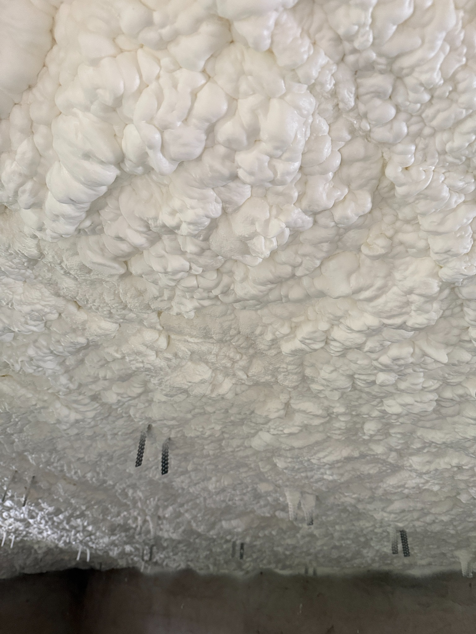 Piana poliuretanowa na suficie, widoczne metalowe elementy mocujące. Biała, nierówna powierzchnia izolacji termicznej. Detal ocieplenia poddasza.