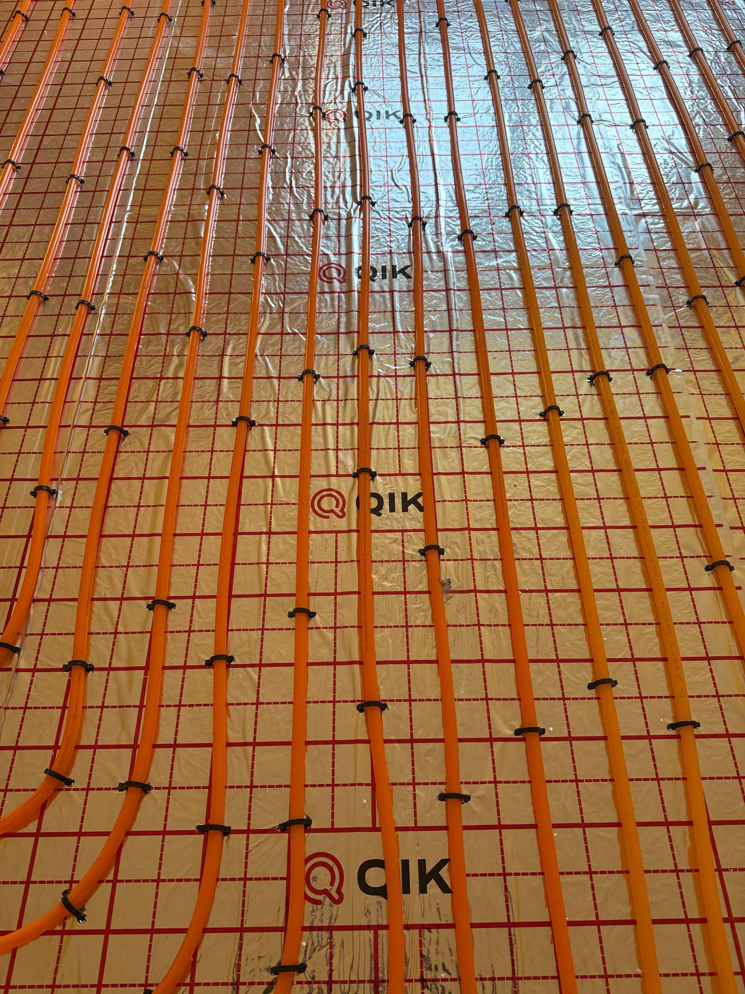 Instalacja ogrzewania podłogowego w Zduńskiej Woli: pomarańczowe rurki równolegle ułożone na srebrnej folii z czerwoną siatką, przymocowane czarnymi uchwytami.