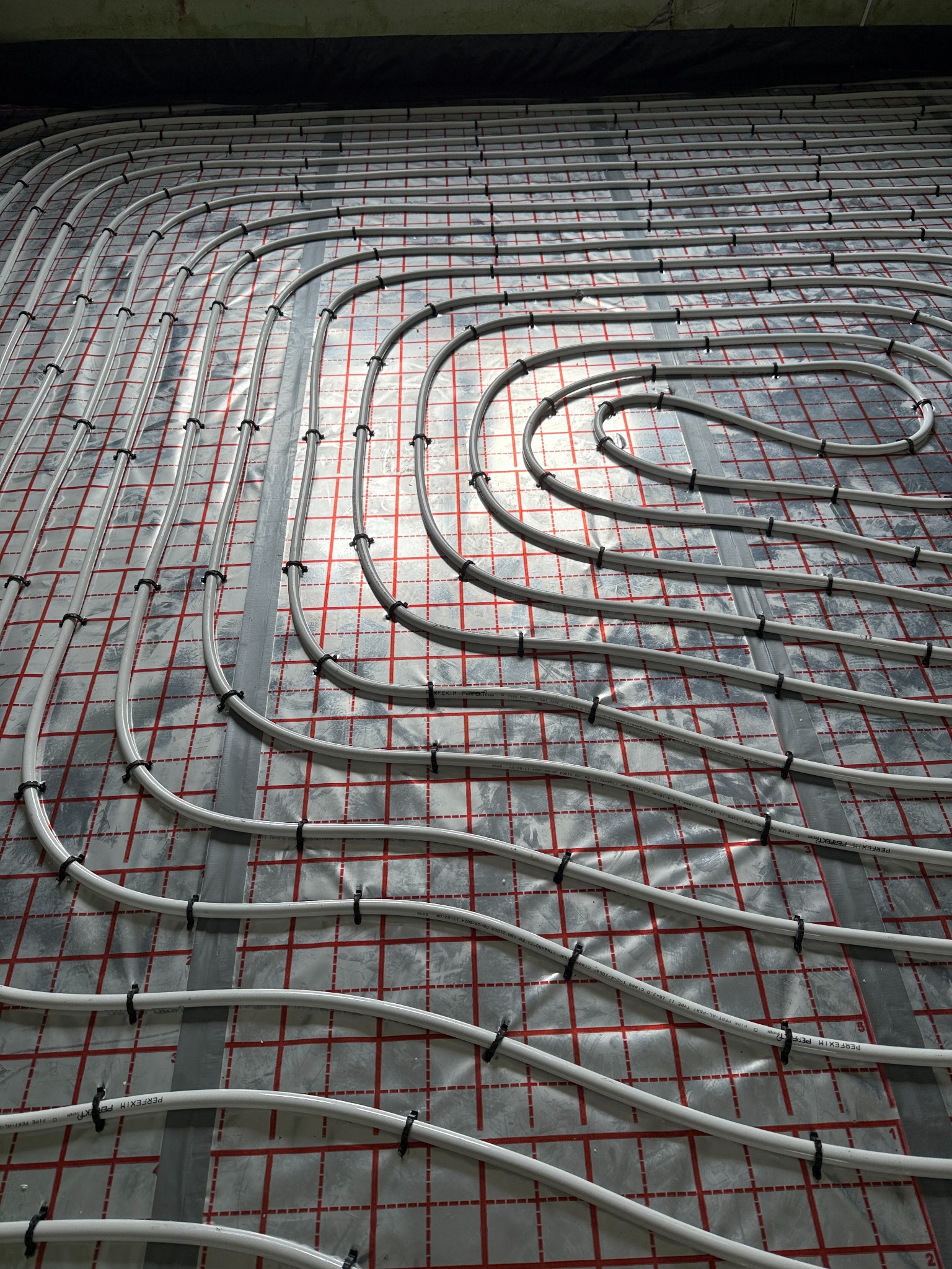 Ułożone, białe rury ogrzewania podłogowego na folii z czerwoną siatką, przymocowane czarnymi uchwytami. Widok z góry, instalacja przed wylaniem posadzki.