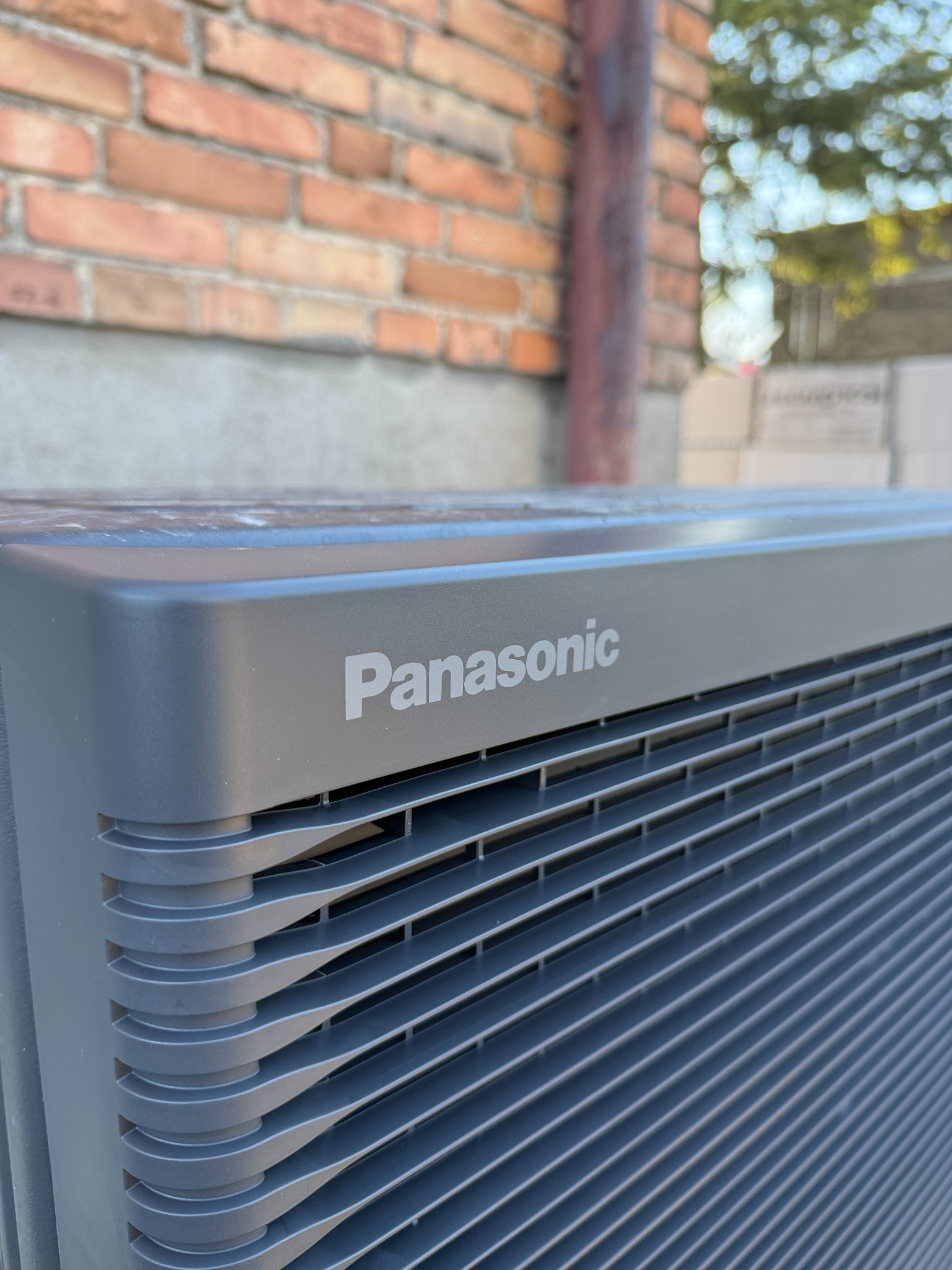 Szary klimatyzator Panasonic na tle ceglanej ściany, widok z dołu, ukazujący detale obudowy i logo, z elementami zieleni w tle.