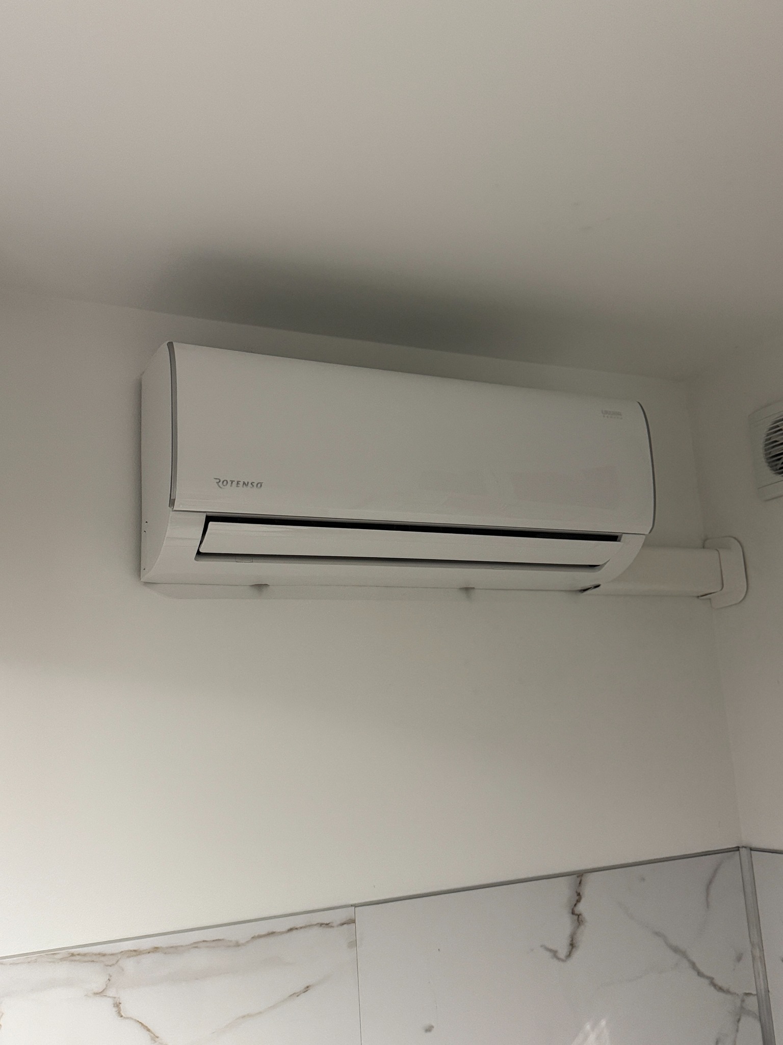 Biały klimatyzator Rotenso zamontowany na ścianie w pomieszczeniu z marmurowymi płytkami. Widoczna instalacja odprowadzająca.