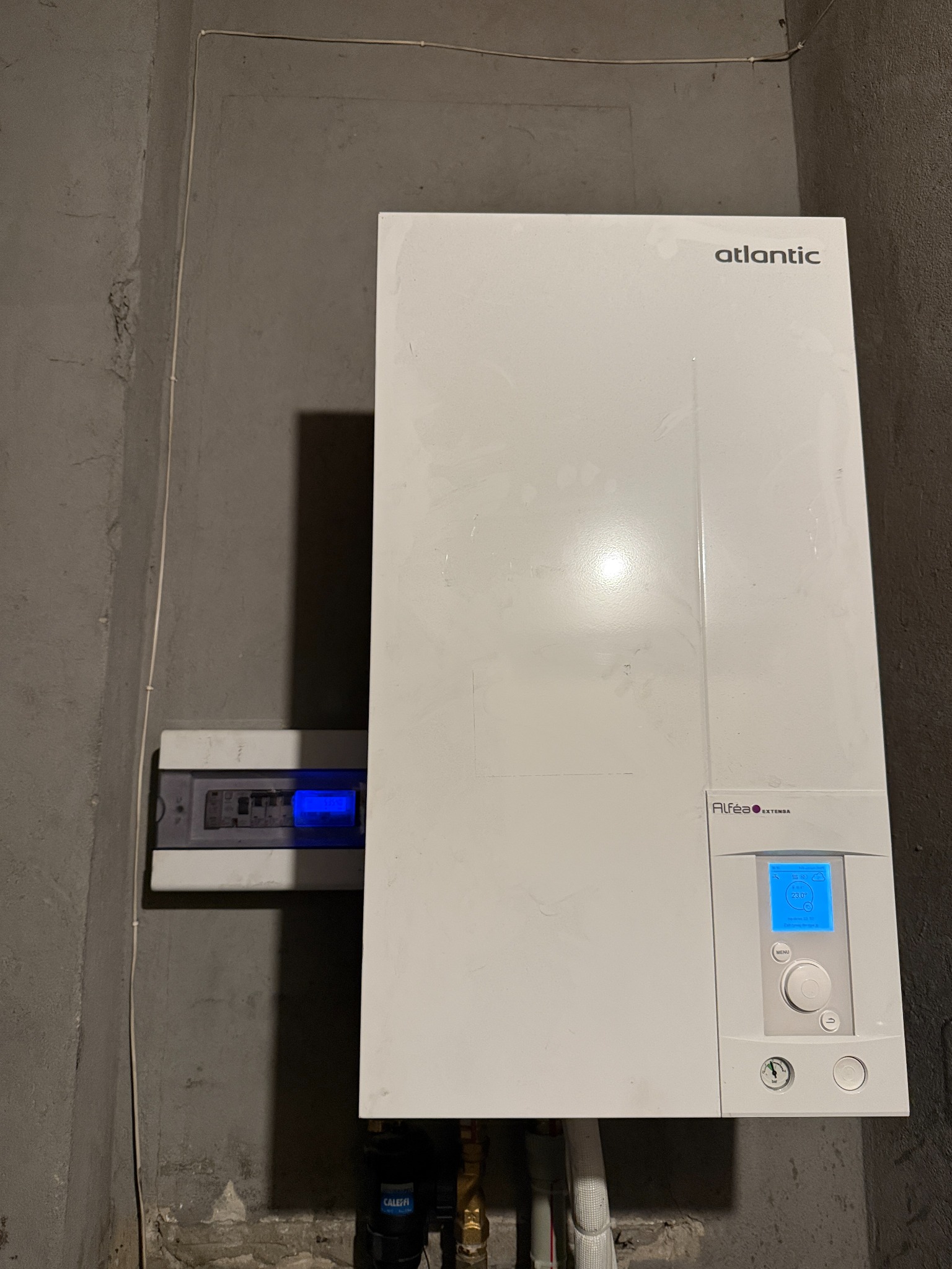 Biały, wiszący piec gazowy Atlantic Alféa Extensa na tle szarej, surowej ściany. Obok skrzynka z bezpiecznikami z niebieskim wyświetlaczem. Widoczne rury i elementy instalacji.