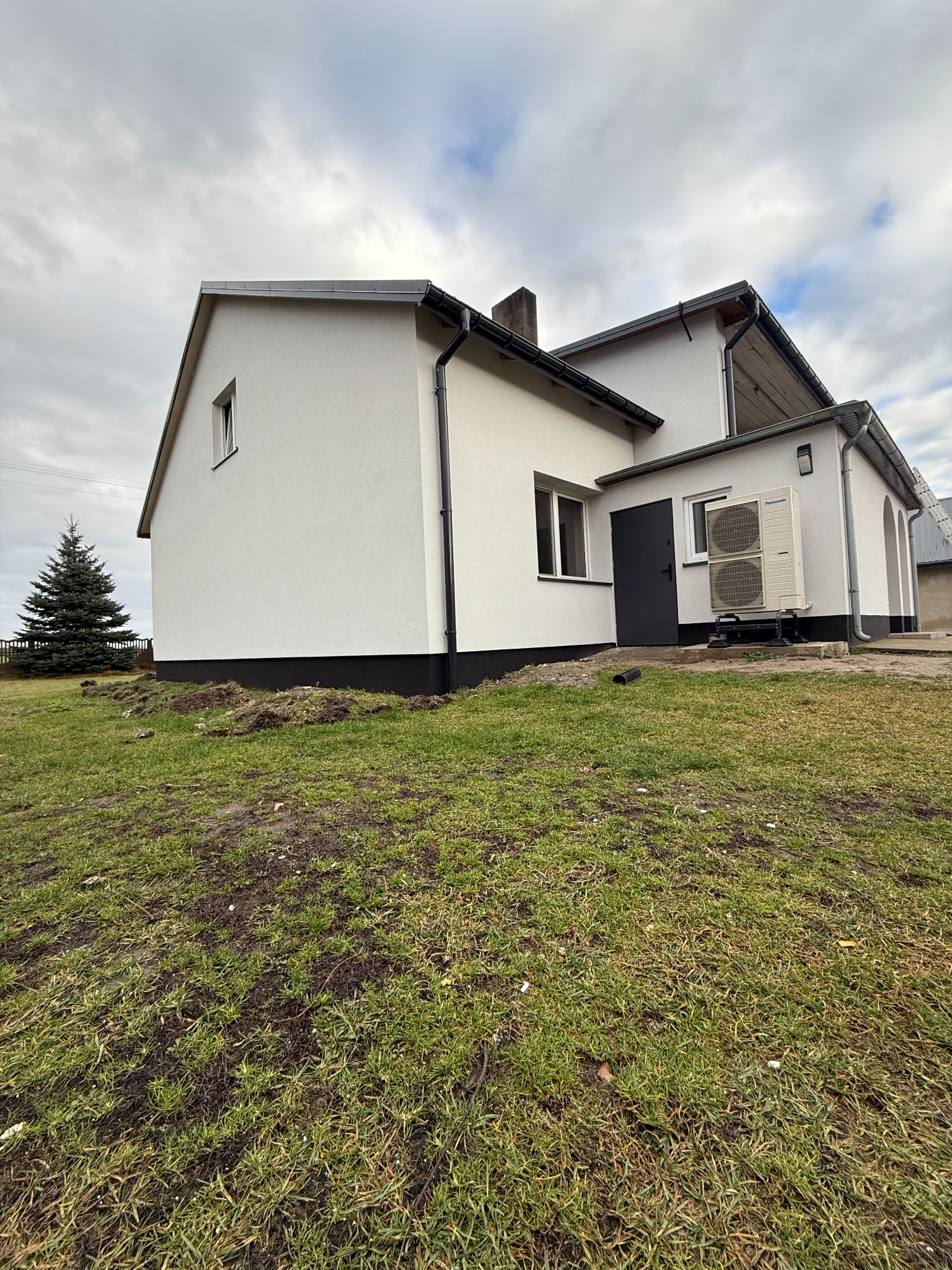 Biały dom z szarą stolarką i pompą ciepła na zewnątrz, widok z trawnika. Świerk w tle, pochmurne niebo.