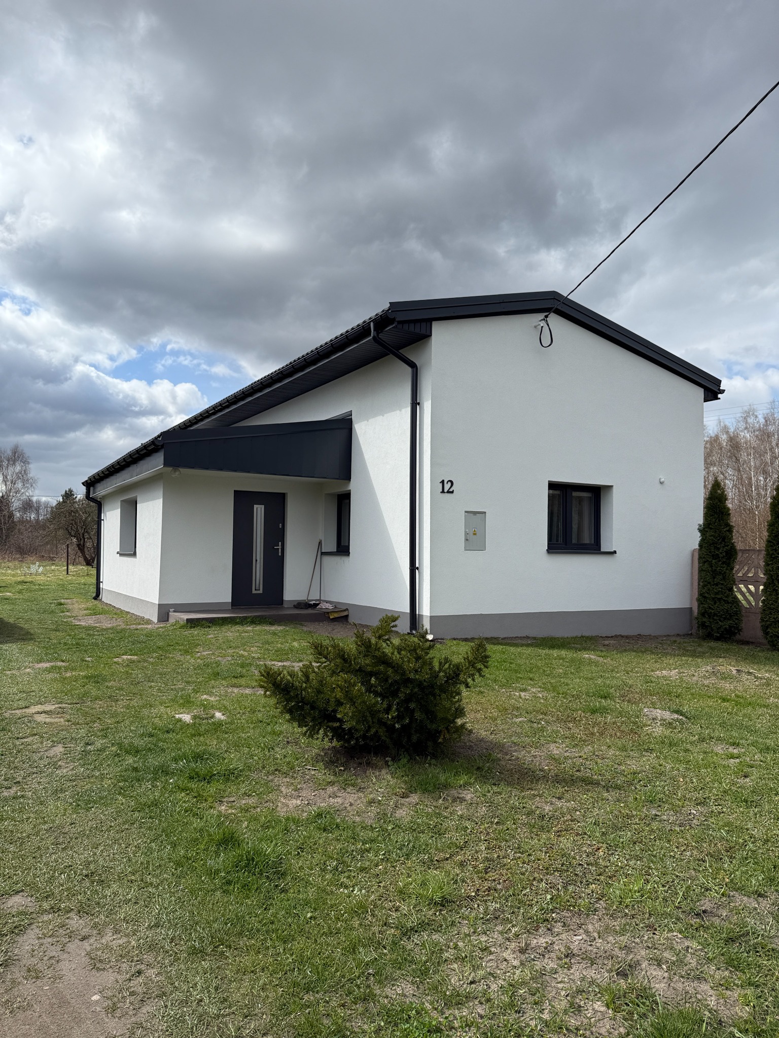 Biały dom z numerem 12, ciemnymi oknami i dachem, otoczony zielonym trawnikiem i krzewami, pod pochmurnym niebem. Widoczna skrzynka elektryczna na elewacji.