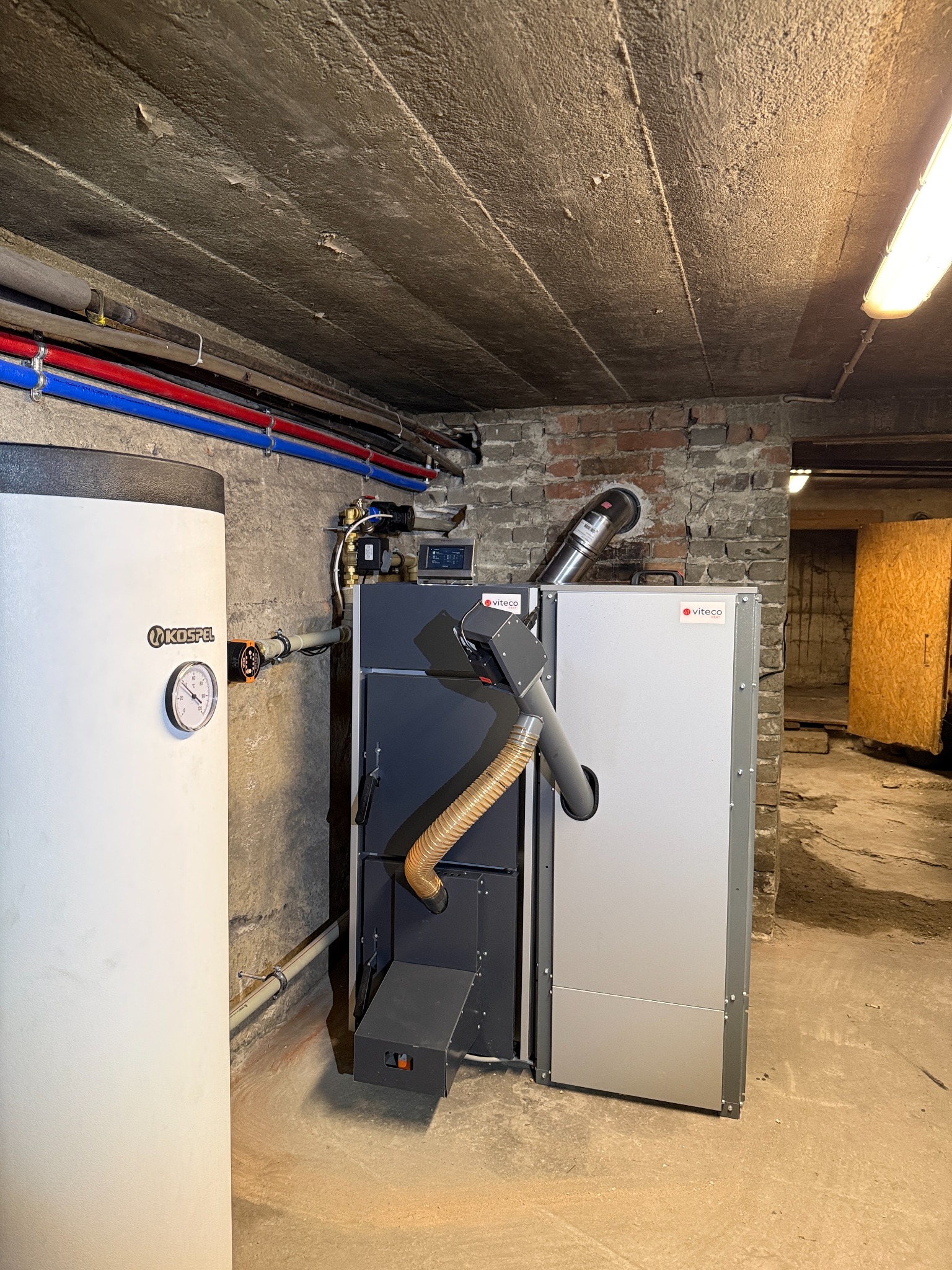 Nowoczesny piec na paliwo stałe Viteco w piwnicy w Zduńskiej Woli. Widoczny zbiornik Kospel, rury instalacji CO i fragment ceglanej ściany.