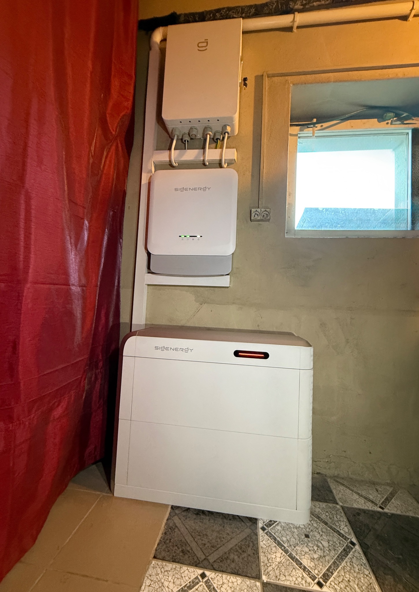 Biały magazyn energii Sigenenergy zamontowany na ścianie w pomieszczeniu z oknem i zasłoną. Widoczne kable i instalacja elektryczna.