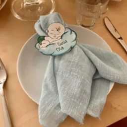 Uroczyste nakrycie stołu z błękitną serwetką, na której umieszczono ozdobny, personalizowany pierścień w kształcie chmurki z rysunkiem śpiącego dziecka i napisem 'Ciocia Ola'.