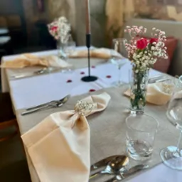 Elegancko nakryty stół z beżowymi serwetkami przewiązanymi sznurkiem i drobnymi kwiatami, świecznikiem i wazonem z czerwonymi różami i gipsówką, dekorowany płatkami róż.