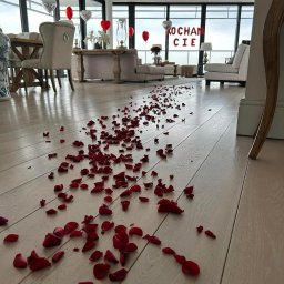 (nie)prostyevent - Romantyczna aranżacja wnętrza: płatki czerwonych róż rozsypane na jasnej podłodze prowadzące do salonu z balonami w kształcie serc i napisem 'KOCHAM CIE'.