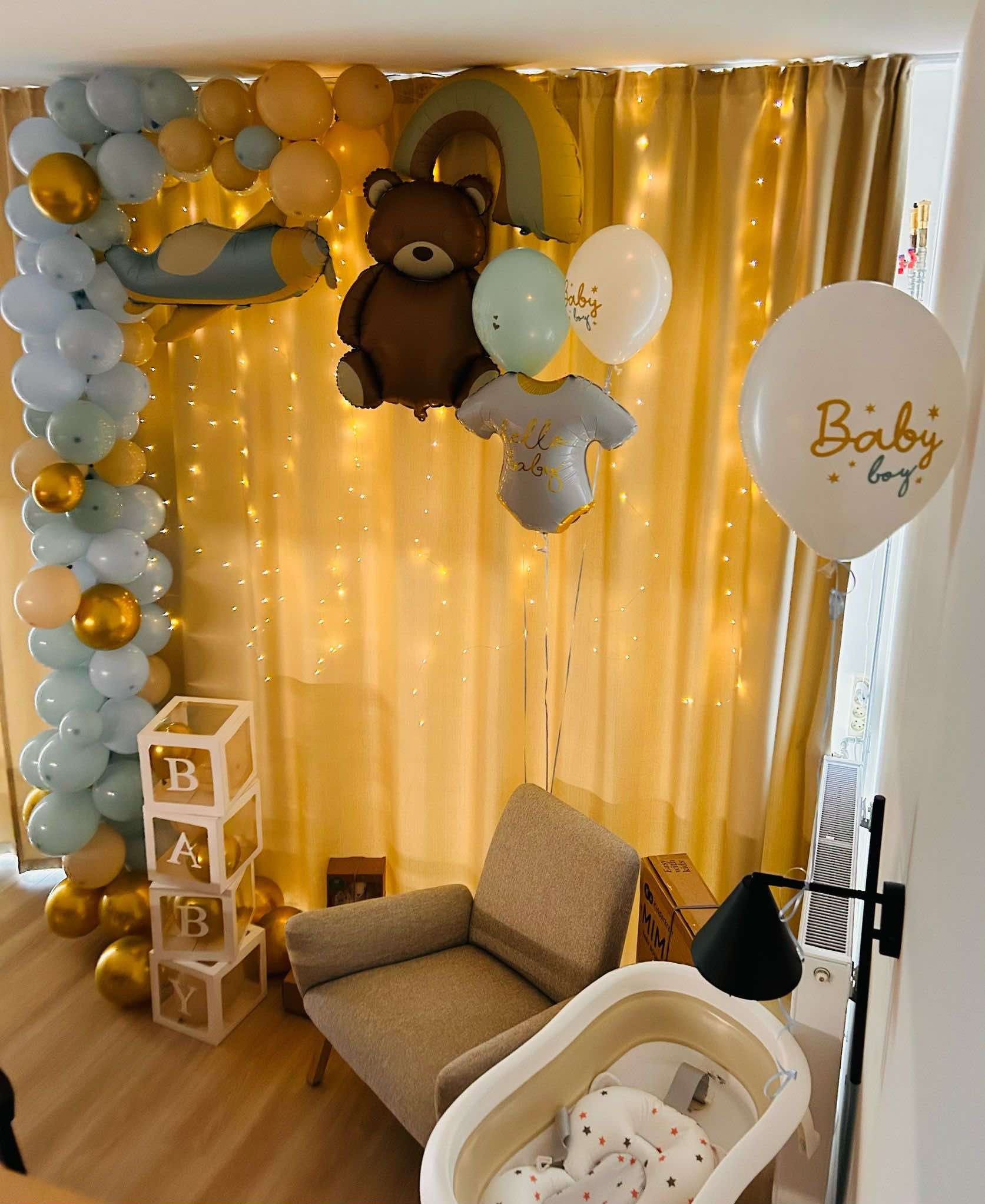 Dekoracja balonowa na baby shower z motywem misia, samolotu i tęczy, podświetlona lampkami, z drewnianymi kostkami z literami 'BABY' i dziecięcą wanienką w tle.