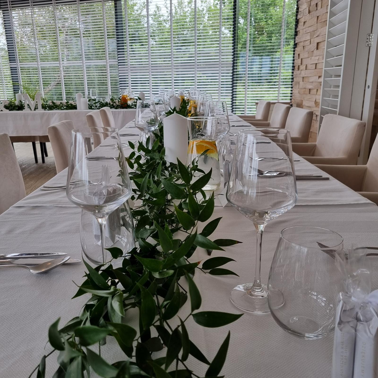 Uroczysta aranżacja stołu z białym obrusem, zielonym girlandą, wysokimi kieliszkami, świecami i dzbankiem z wodą i pomarańczą, w tle okno z żaluzjami i ściana z kamienia.