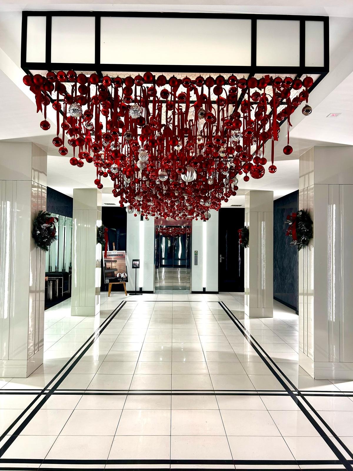 Eleganckie lobby hotelowe udekorowane na święta Bożego Narodzenia, z dużą instalacją z czerwonych i srebrnych bombek zawieszonych pod sufitem oraz wieńcami na kolumnach.