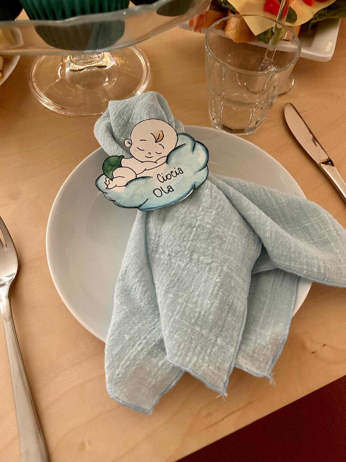 Uroczyste nakrycie stołu z błękitną serwetką, na której umieszczono ozdobny, personalizowany pierścień w kształcie chmurki z rysunkiem śpiącego dziecka i napisem 'Ciocia Ola'.