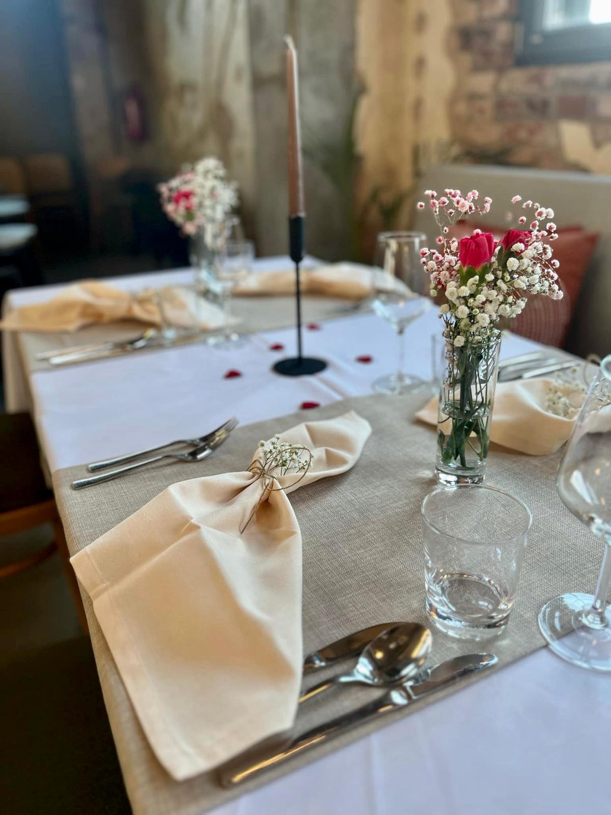 Elegancko nakryty stół z beżowymi serwetkami przewiązanymi sznurkiem i drobnymi kwiatami, świecznikiem i wazonem z czerwonymi różami i gipsówką, dekorowany płatkami róż.